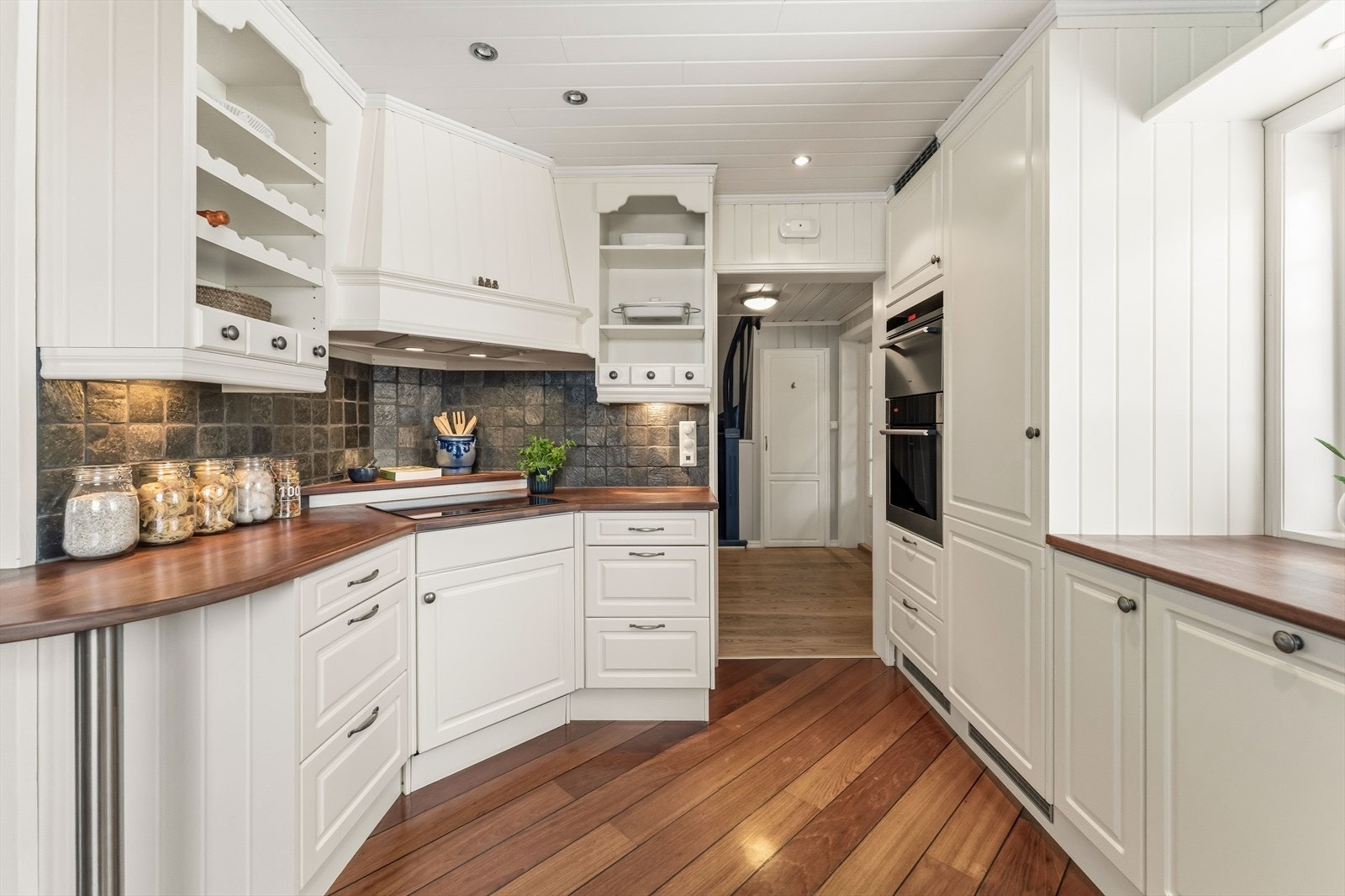 Innredningen har et klassisk uttrykk med hvite profilfronter, åpne hyller, vitrineskap og heltre benkeplater i en fargetone som harmonerer med gulvet. Galleribilde