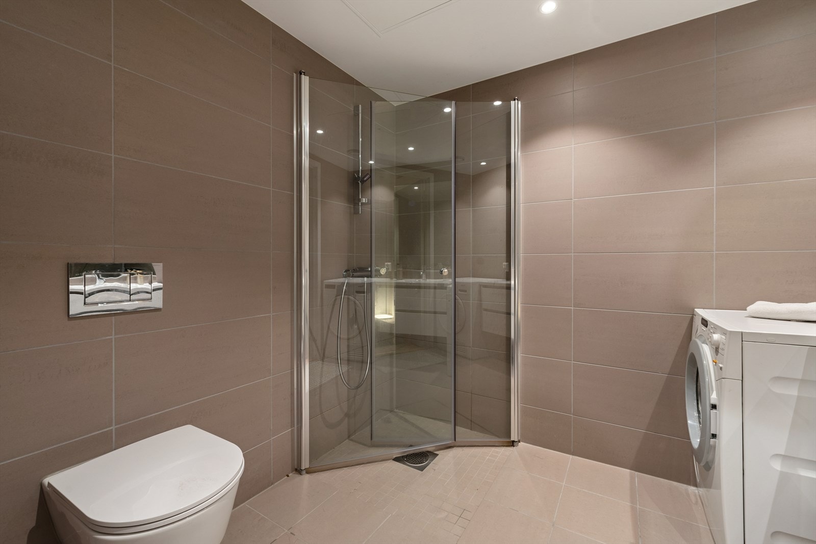 Rommet er innredet med servant med slette fronter, veggmontert toalett og dusjhjørne med to svingbare glassdører. Galleribilde