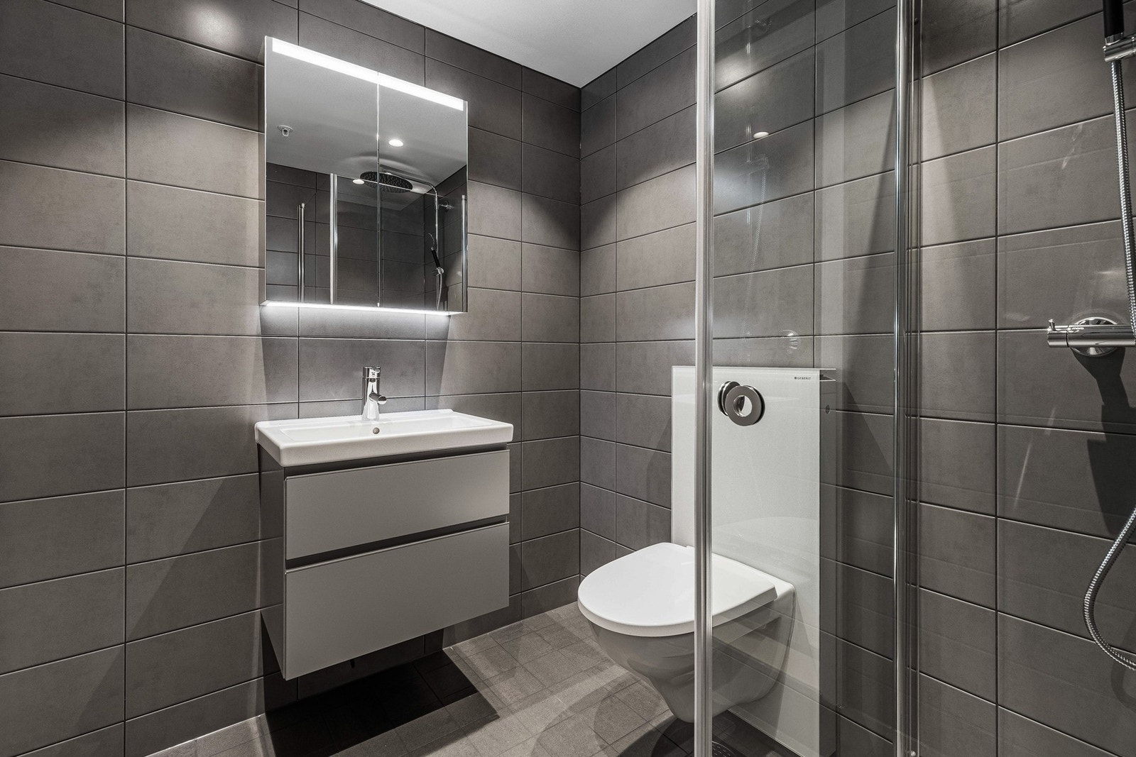 Her er det mørkegrå fliser på veggene, downlights i himlingen og grå fliser på gulv med gulvvarme. Baderomsinnredningen har grå, glatte fronter og består av en heldekkende servant på en skuffeseksjon og et dobbelt speilskap med integrert belysning. Galleribilde