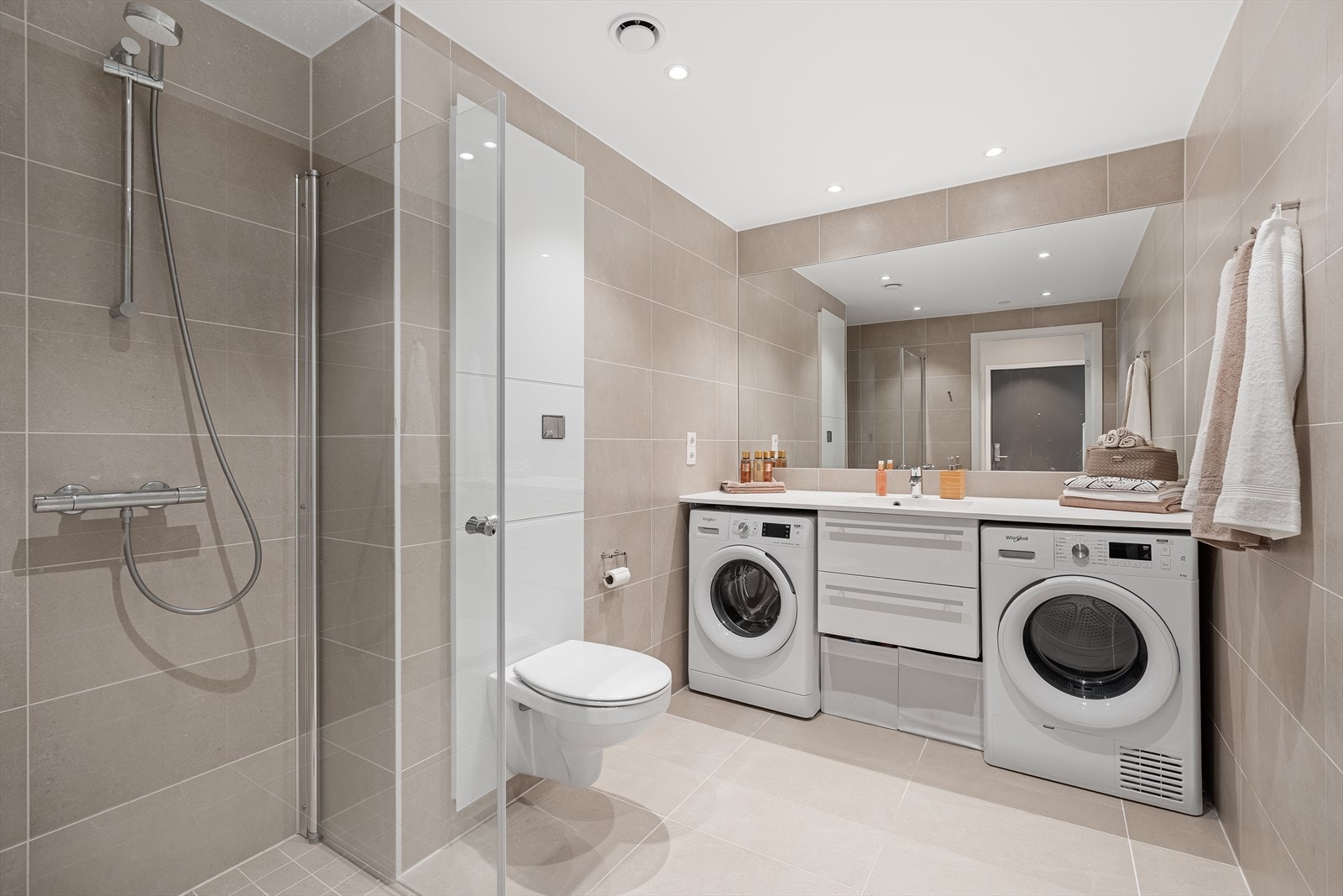 Badet er helfliset med varme i gulvet og inneholder en praktisk løsning med baderomsinnredning med plass til vaskemaskin og tørketrommel, vegghengt toalett, samt dusjnisje med innfellbare dusjdører. Galleribilde