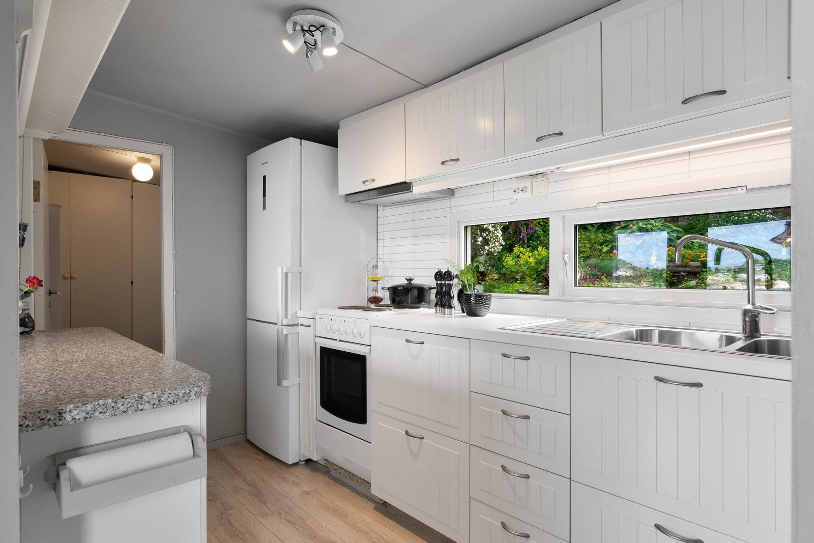 Kjøkkeninnredningen er montert langs to vegger og har hvite, profilerte fronter og benkeplate i laminat. Det er nedfelt oppvaskkum med avrenningsfelt i den ene benken. Galleribilde