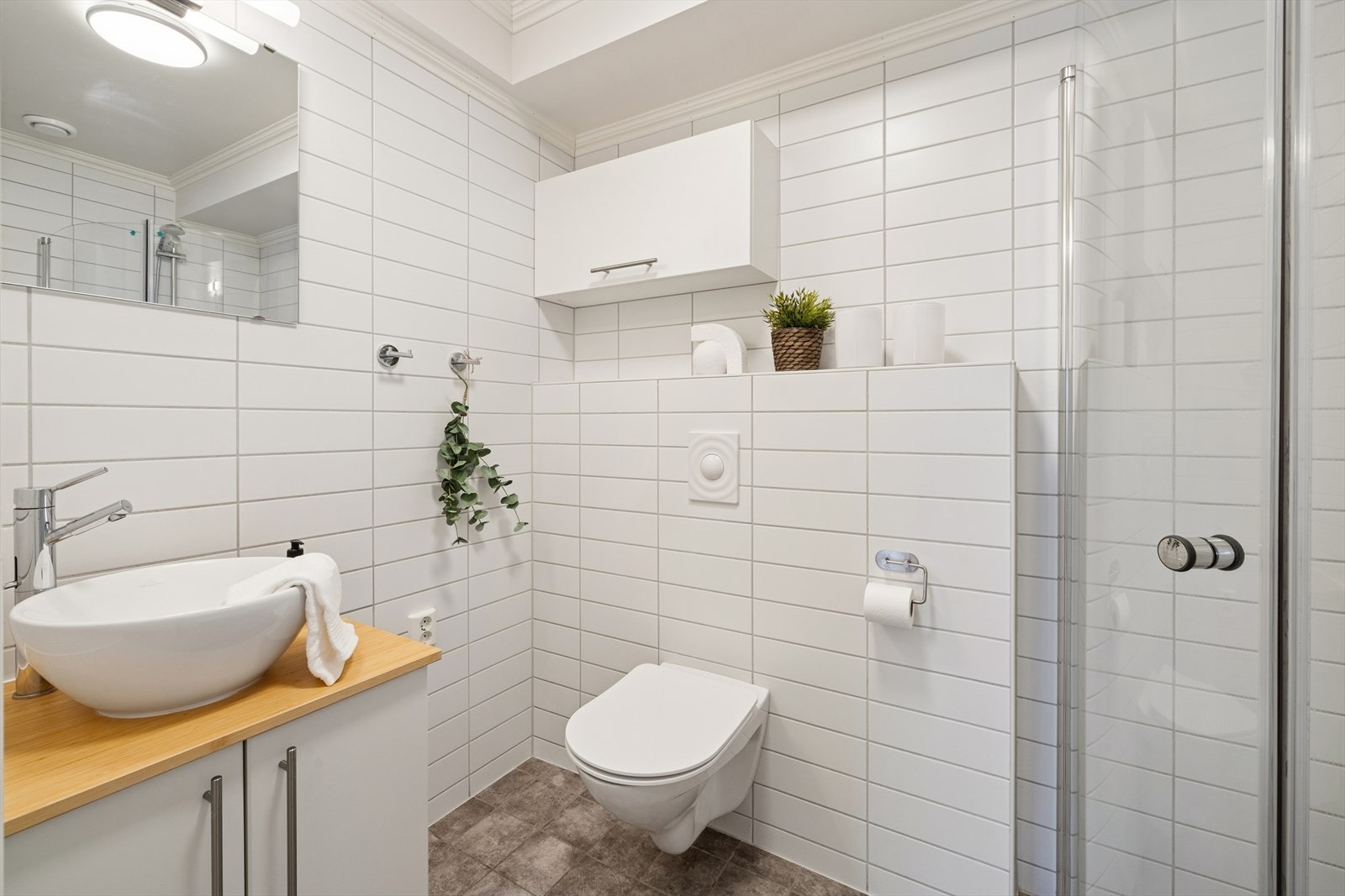 Badet i 1. etasje har flislagte overflater og elektriske varmekabler i gulv. Innredningen består av et benkeskap og veggskap, toppmontert servant og speil. I tillegg har badet et veggmontert toalett og en dusj med innfellbare vegger. Galleribilde