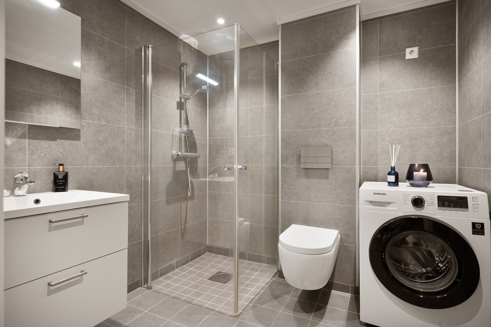 Moderne flislagt bad med gulvvarme, dusjhjørne, opplegg for vaskemaskin, servant og vegghengt toalett. Galleribilde