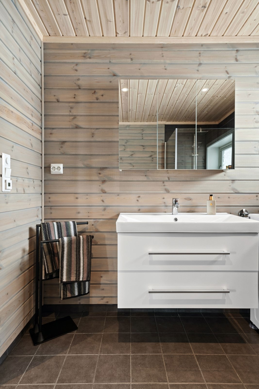 Moderne baderomsinnredning med skuffer og heldekkende servant, vegghengt speilskap montert over. Baderommet er også utstyrt med dusjhjørne med innfellbare glassdører og både hånd- og regnfallsdusj, gulvmontert toalett og opplegg for vaskemaskin. Galleribilde