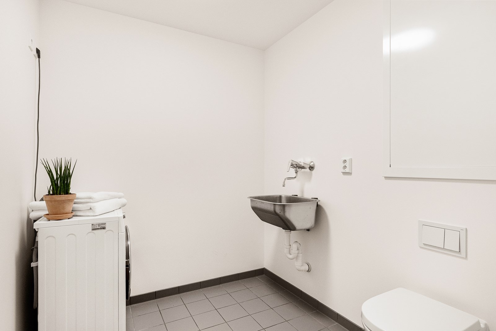 Praktisk og funksjonelt vaskerom med flislagt gulv og varmekabler. Rommet har malte, slette flater på vegger og i tak, noe som gir et lyst og ryddig uttrykk. Vaskerommet er innredet med utslagsvask, toalett, samt opplegg og god plass til vaskemaskin. Galleribilde