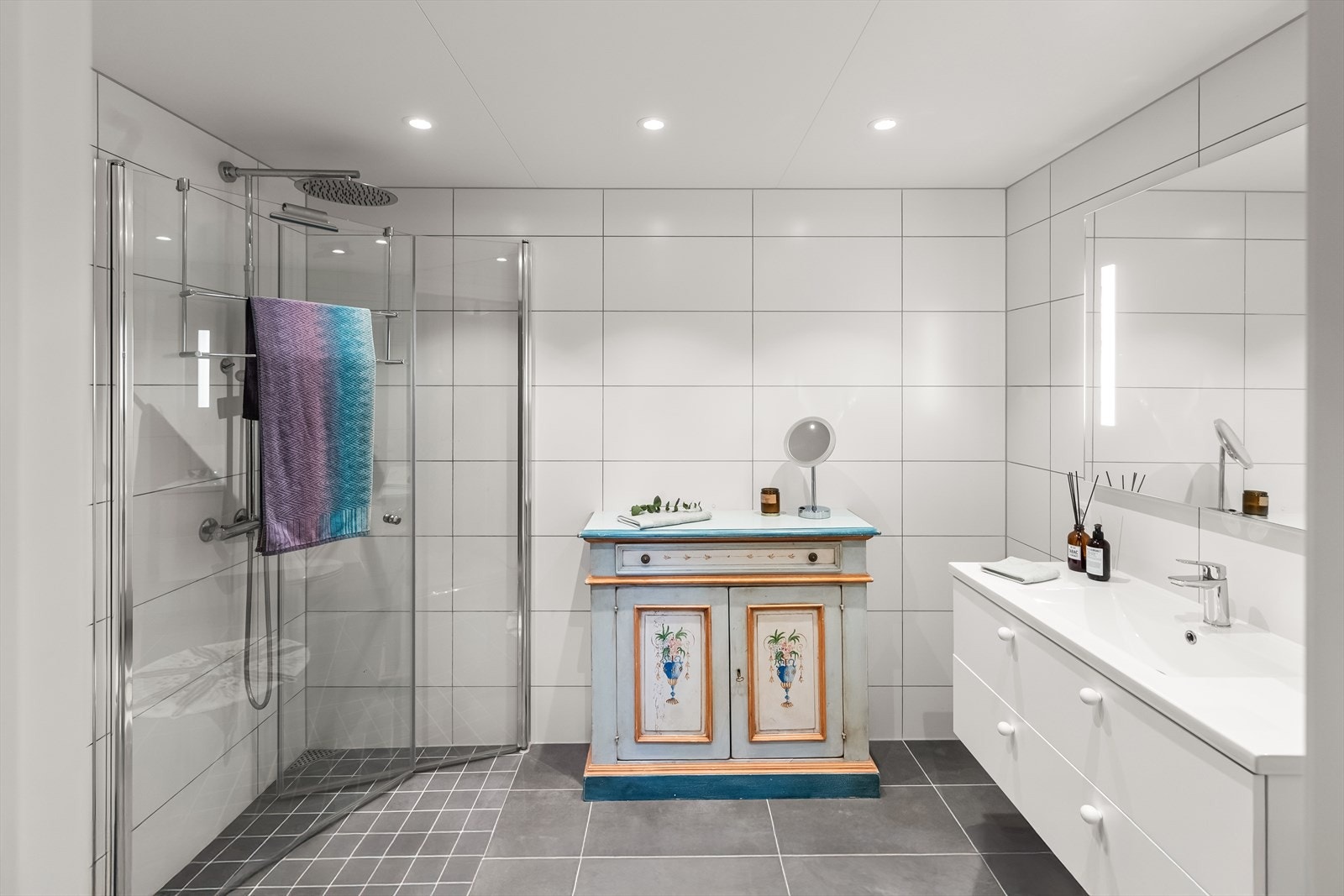 På en hvit skuffeinnredning er det en heldekkende servant, og over er det et stort speil med integrert belysning. Installert regndusjhoder . Galleribilde