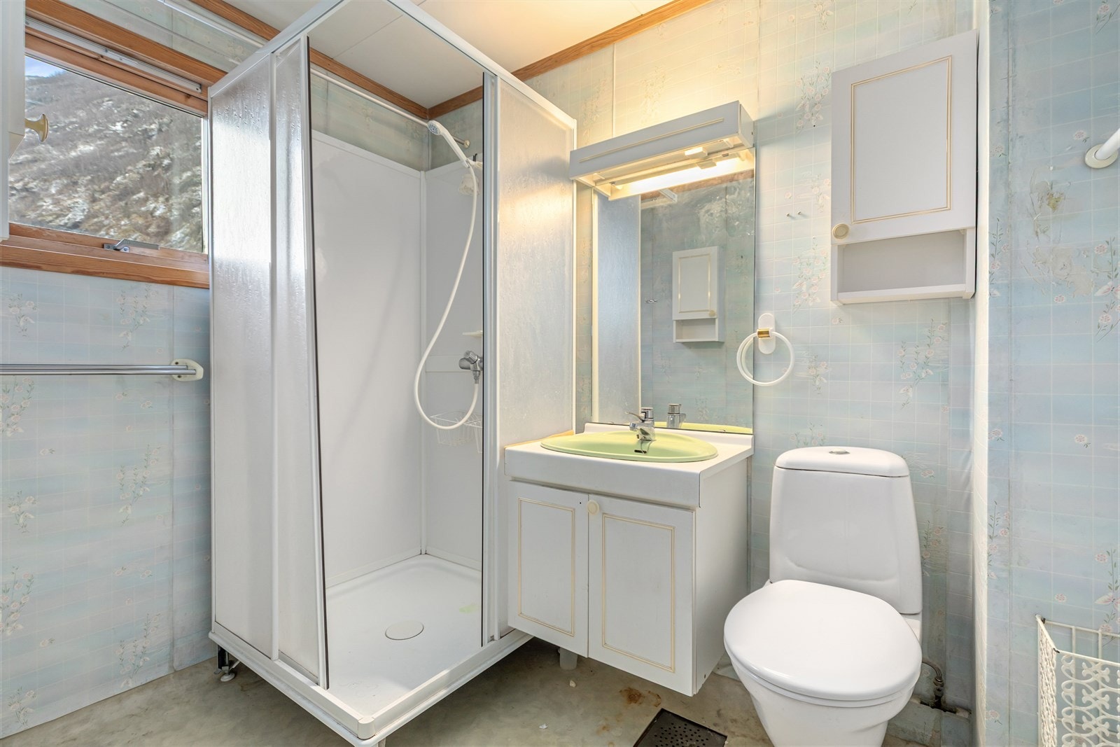 Bad av eldre stand utstyrt med dusjhjørne, toalett og servant med innredning. Galleribilde