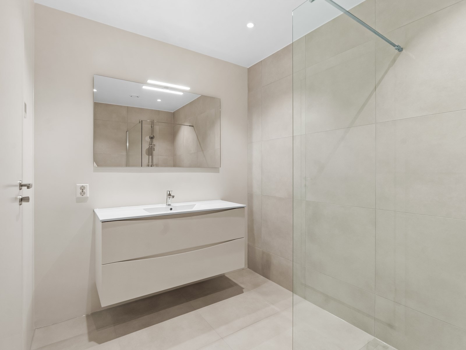 Moderne og delikat bad med fliser på gulv og vegger. Baderomsinnredningen består av servant, dusjhjørne, toalett og et speil med belysning. Galleribilde