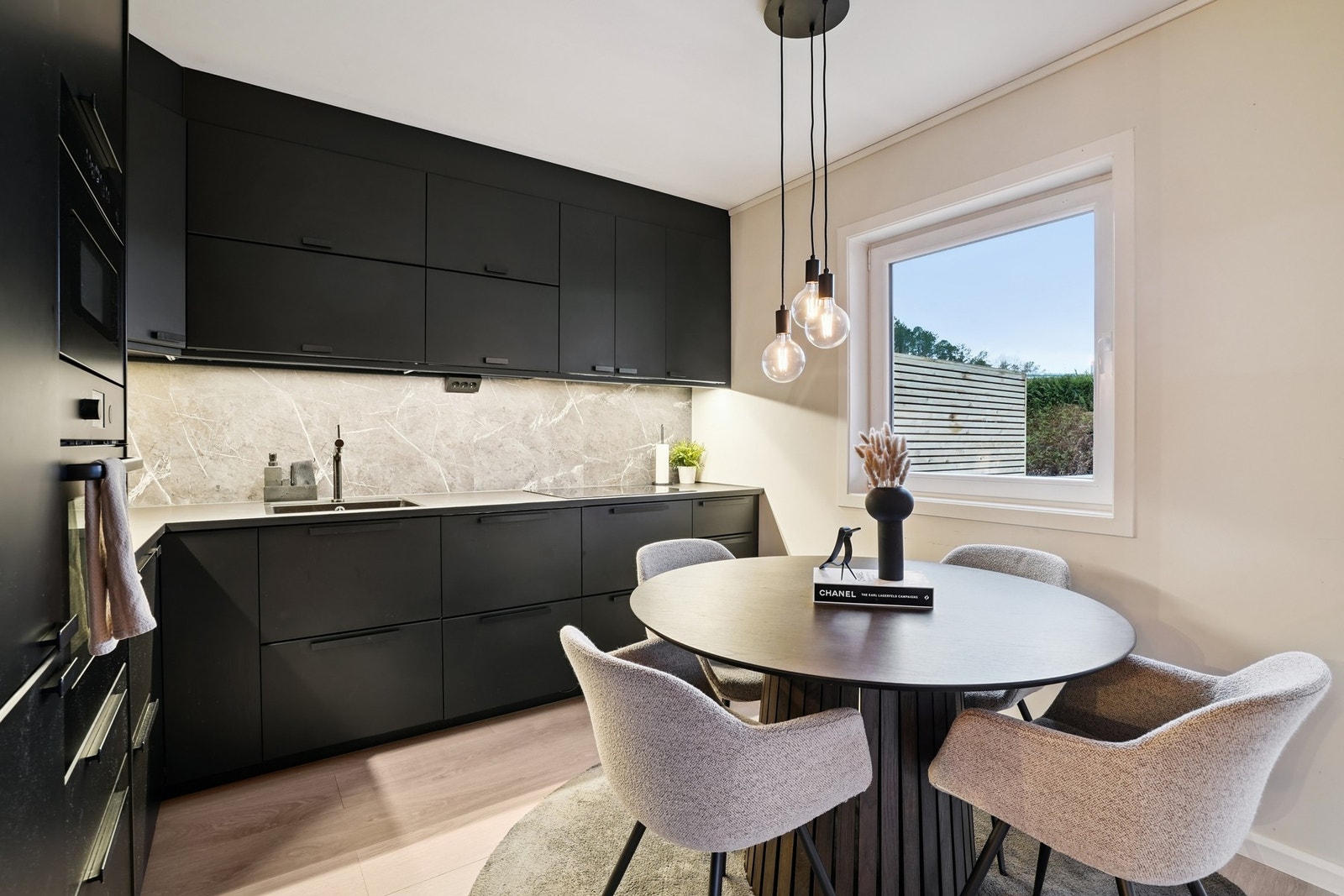 Stilren spisegruppe plassert ved vinduet, med myke stoler og behagelig lys som gir kjøkkenet en varm og innbydende atmosfære. Galleribilde