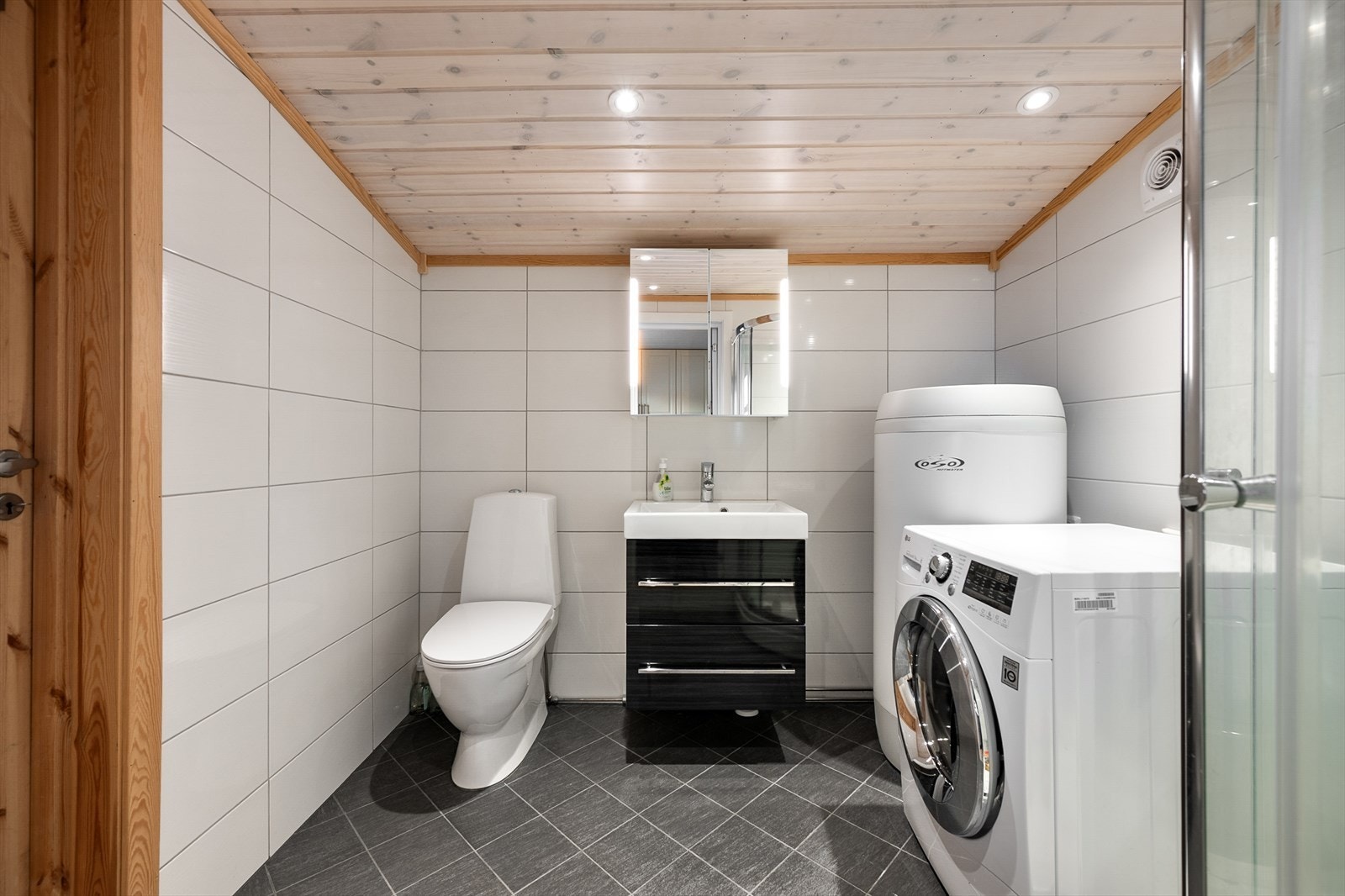 Flislagt bad med vegghengt toalett, dusjhjørne og opplegg for vaskemaskin. Galleribilde