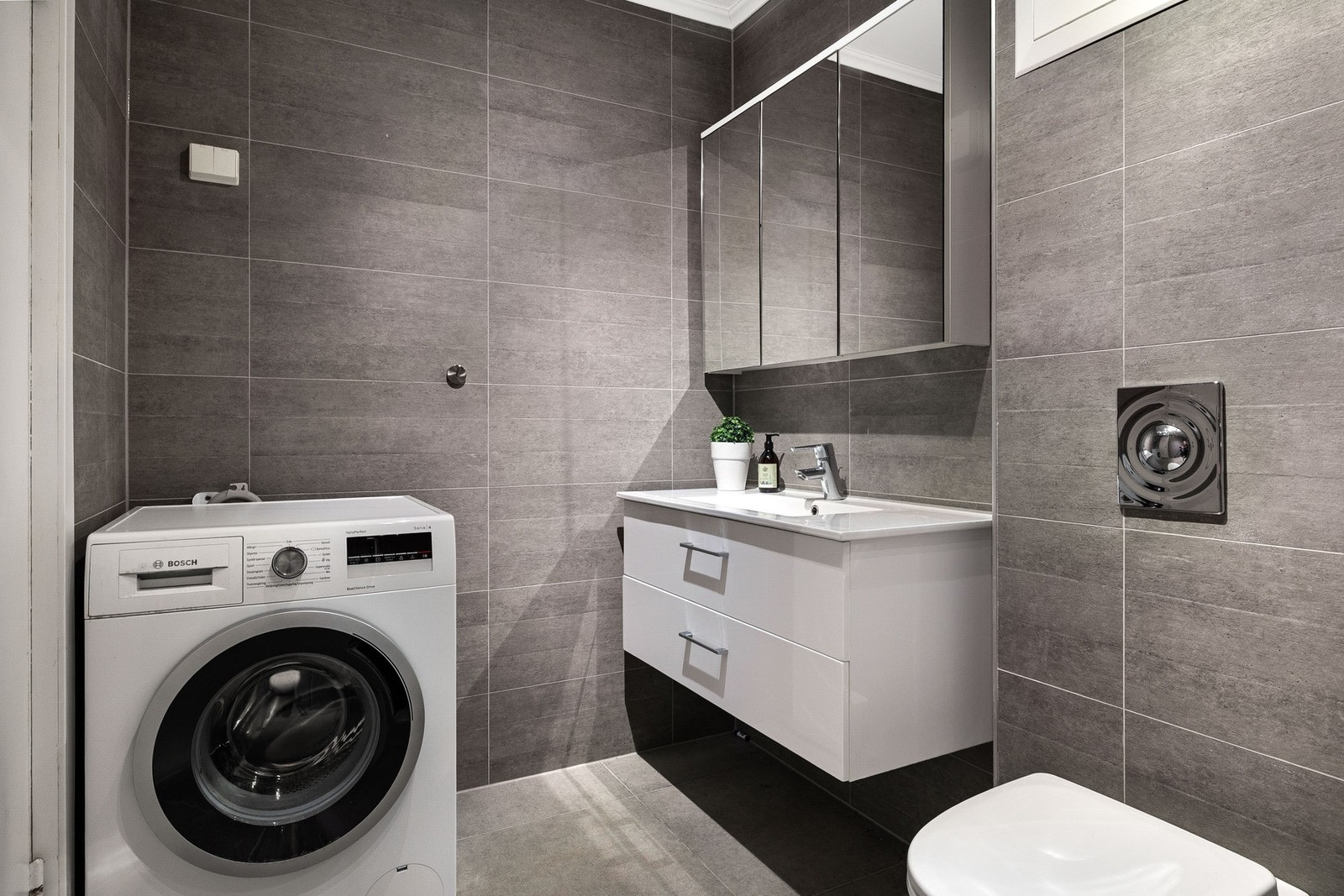 Flislagt bad med varmekabler i gulv og er innredet med veggmontert toalett, servant med innredning, dusjhjørne samt opplegg for vaskemaskin. Galleribilde