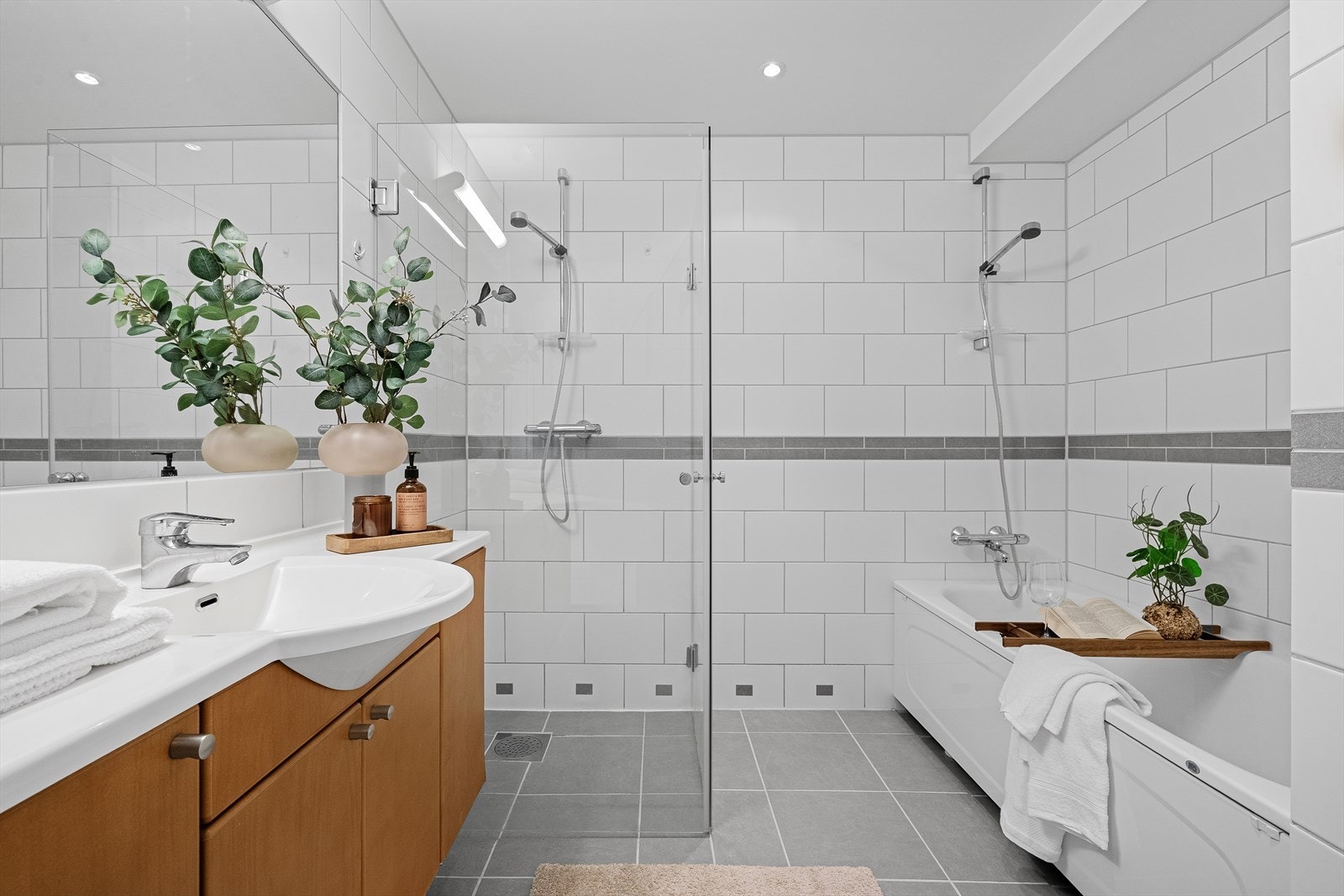 Badet har flislagt gulv, lyse overflater og spotter i taket, og er innredet med badekar, dusjsone, toalett, stort speil og lys baderomsinnredning. Galleribilde