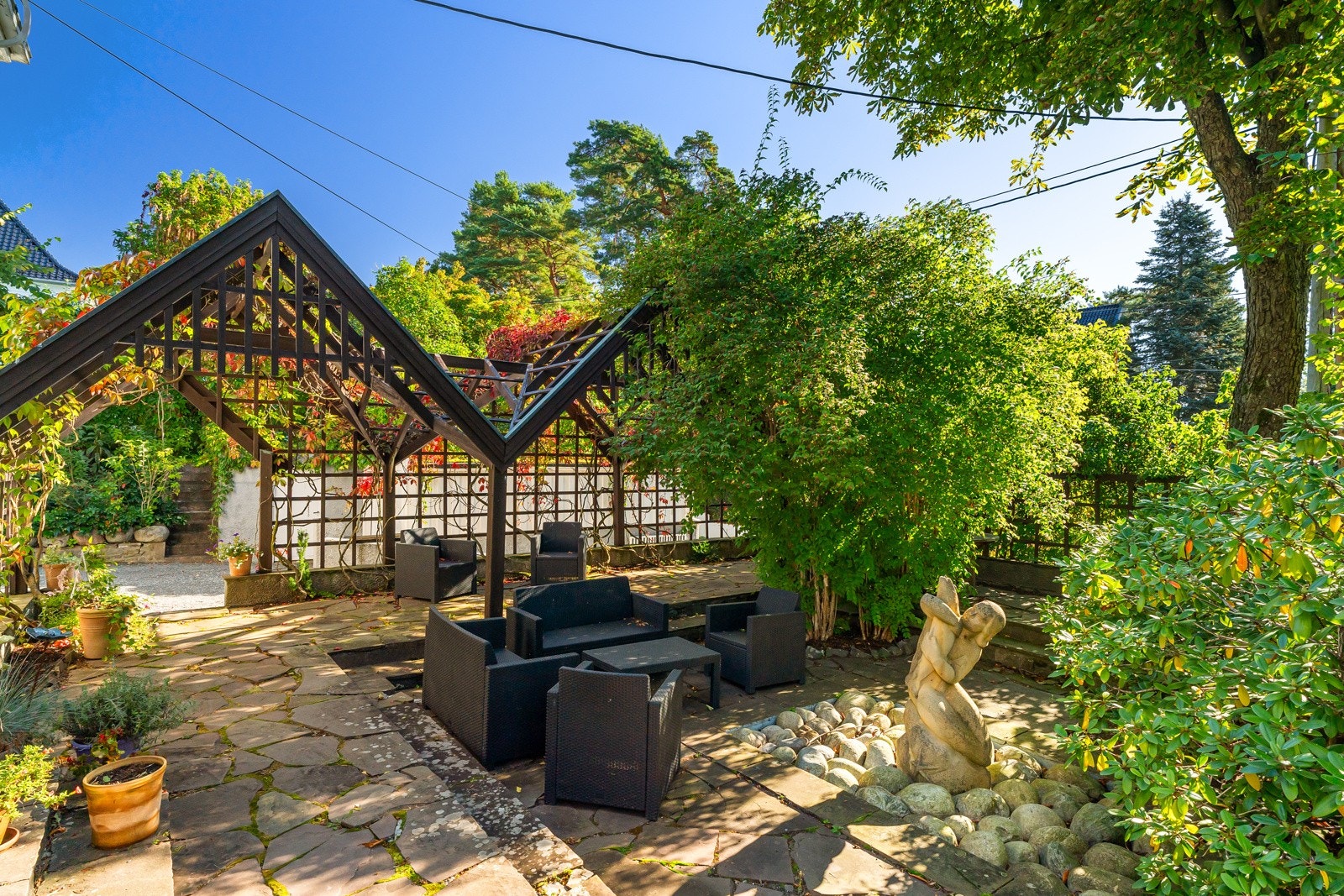 Fra den nedre verandaen er det trapp ned til uteplassen ved pergolaen med hengeplanter og stort dekke av skifer. I tilknytting til uteplassen er det et mindre springvann med en statue av den Islandske artisten Guðmundur fra Miðdal på Island. Galleribilde