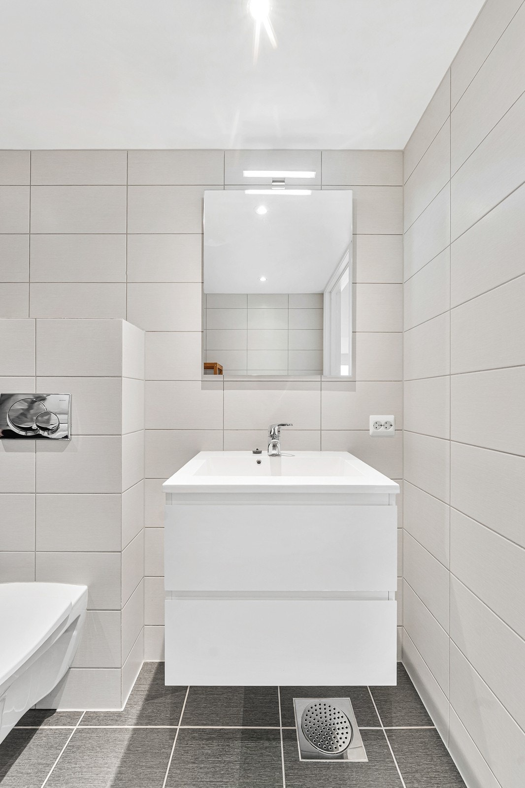 Badet er utstyrt med dusjhjørne med glassvegger, vegghengt toalett, servant med innredning, samt opplegg for vaskemaskin. Galleribilde