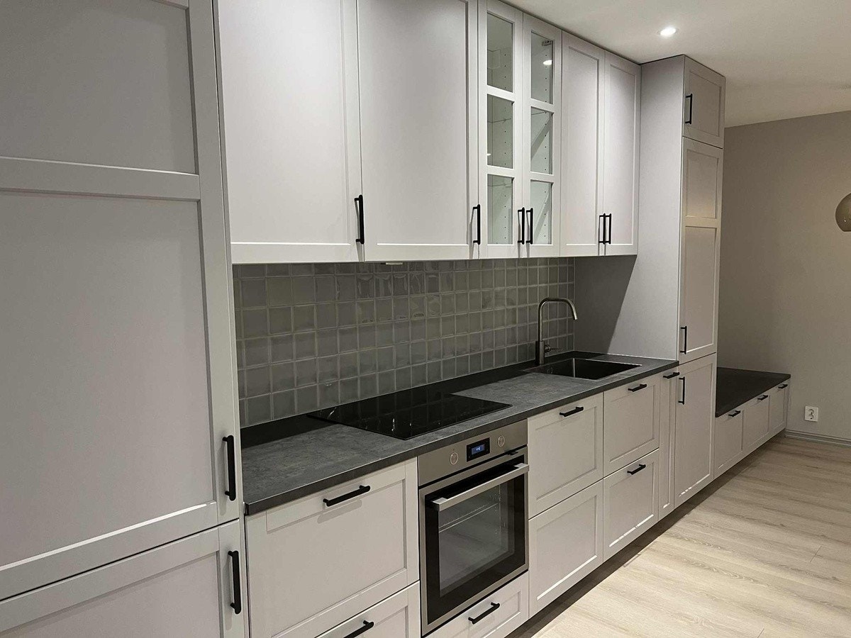 Stort innholdsrikt kjøkken med lys grå profilerte fronter, fliser mellom benk og overskap og integrerte hvitevarer - kjøl/frys, stekeovn, koketopp, ventilator, komfyrvakt og oppvaskmaskin. Galleribilde