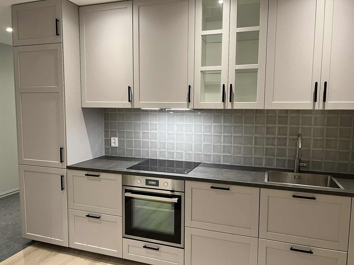 Stort innholdsrikt kjøkken med lys grå profilerte fronter, fliser mellom benk og overskap og integrerte hvitevarer - kjøl/frys, stekeovn, koketopp, ventilator, komfyrvakt og oppvaskmaskin. Galleribilde