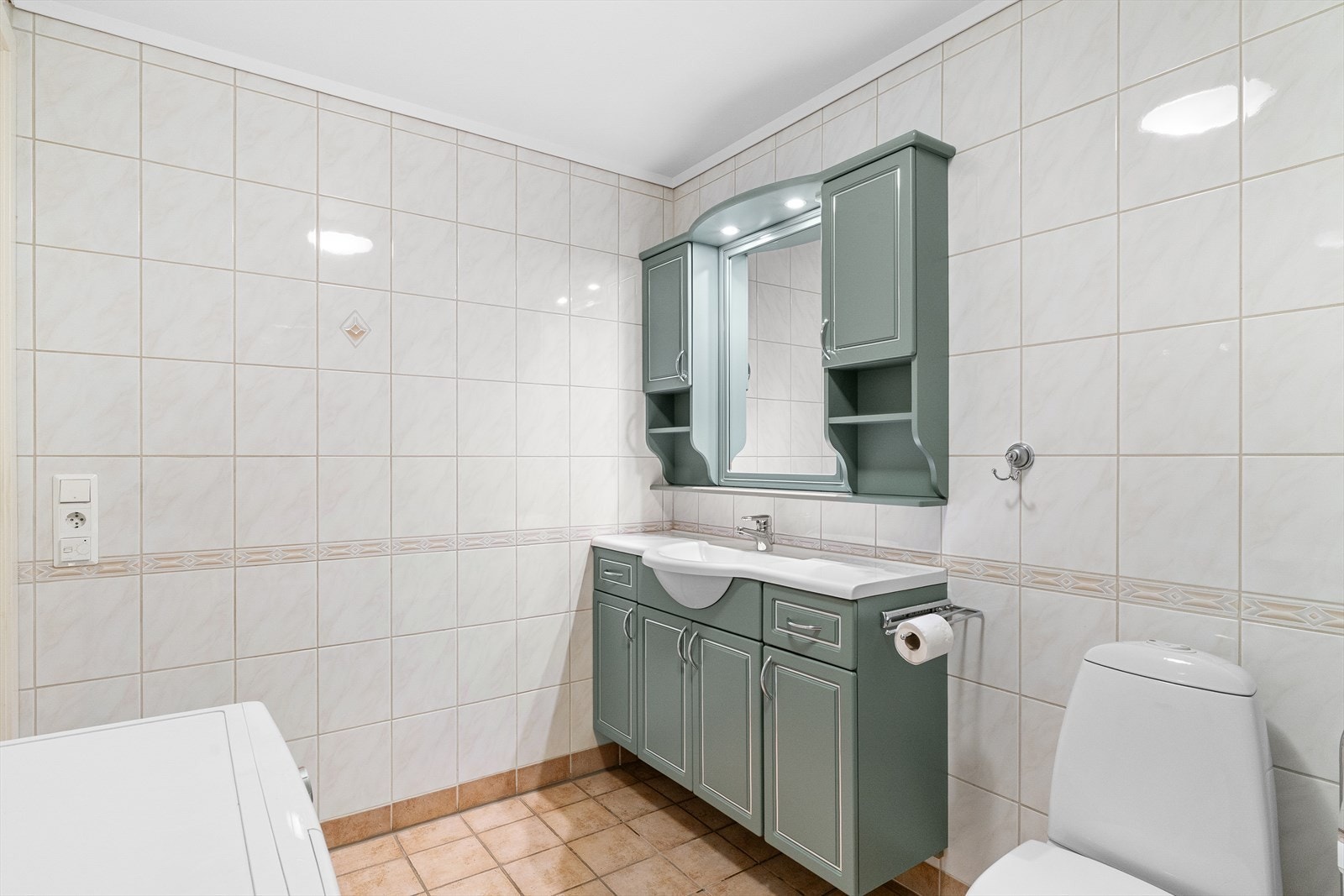 Eldre flislagt bad med varmekabler i gulv. Badet er innredet med nedfelt servant med rom for oppbevaring, speil, toalett og inn fliset badekar med dusjarmatur. Rommet har opplegg for vaskemaskin. Galleribilde