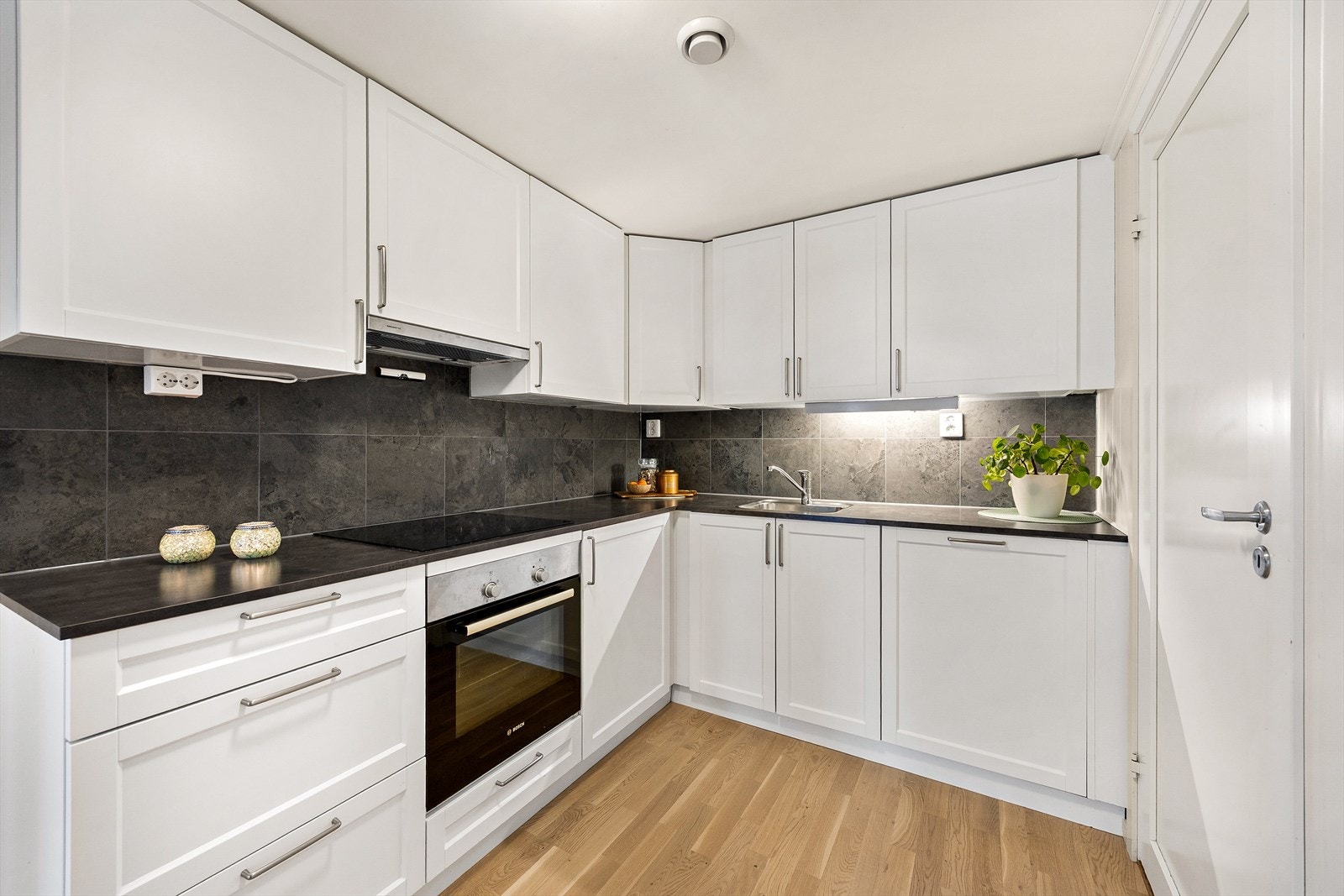 Den vinkelformede og takhøye innredningen har hvite, profilerte fronter og mørk laminatbenkeplate med oppvaskkum i stål. Galleribilde