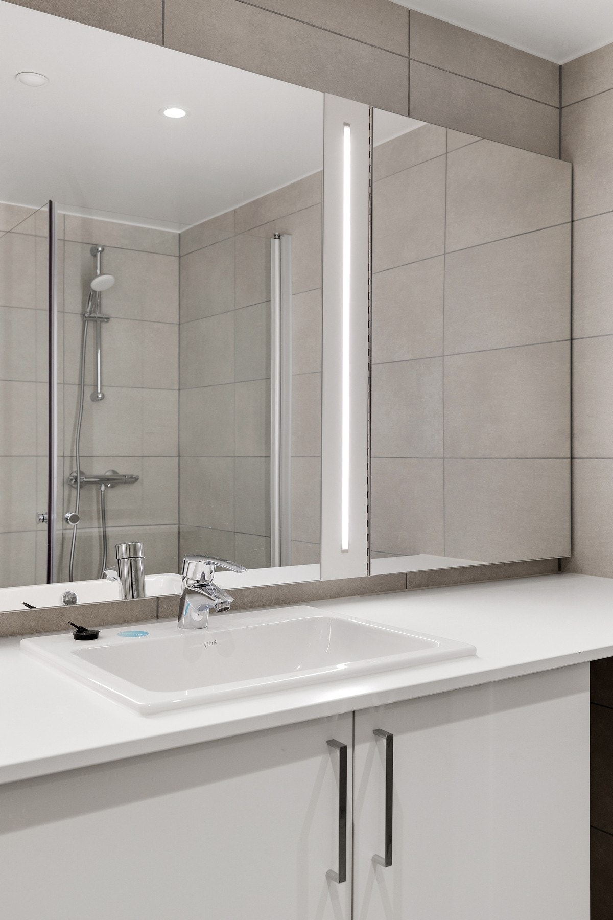 Baderomsinnredningen har glatte, hvite skapfronter og en heldekkende hvit servantplate. Over vasken er det et utvendig speil i samme bredde som innredningen, med integrert belysning. Galleribilde