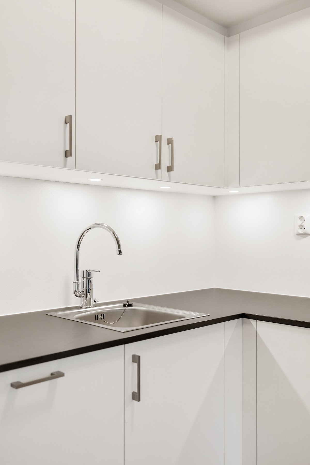 Kjøkkenet har et stilrent design med glatte, hvite fronter og håndtak i rustfritt stål, kombinert med en mørk grå laminat benkeplate. Galleribilde