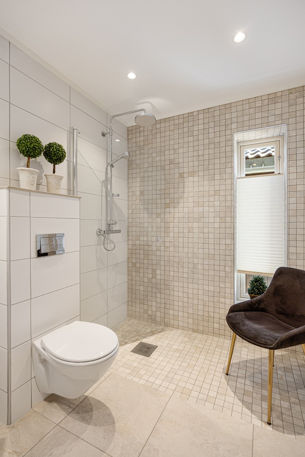 Badene har lysbeige fliser på gulv med gulvvarme, matchende mosaikk på gulv og én vegg i dusjsonen, samt hvite fliser på øvrige veggflater. Galleribilde