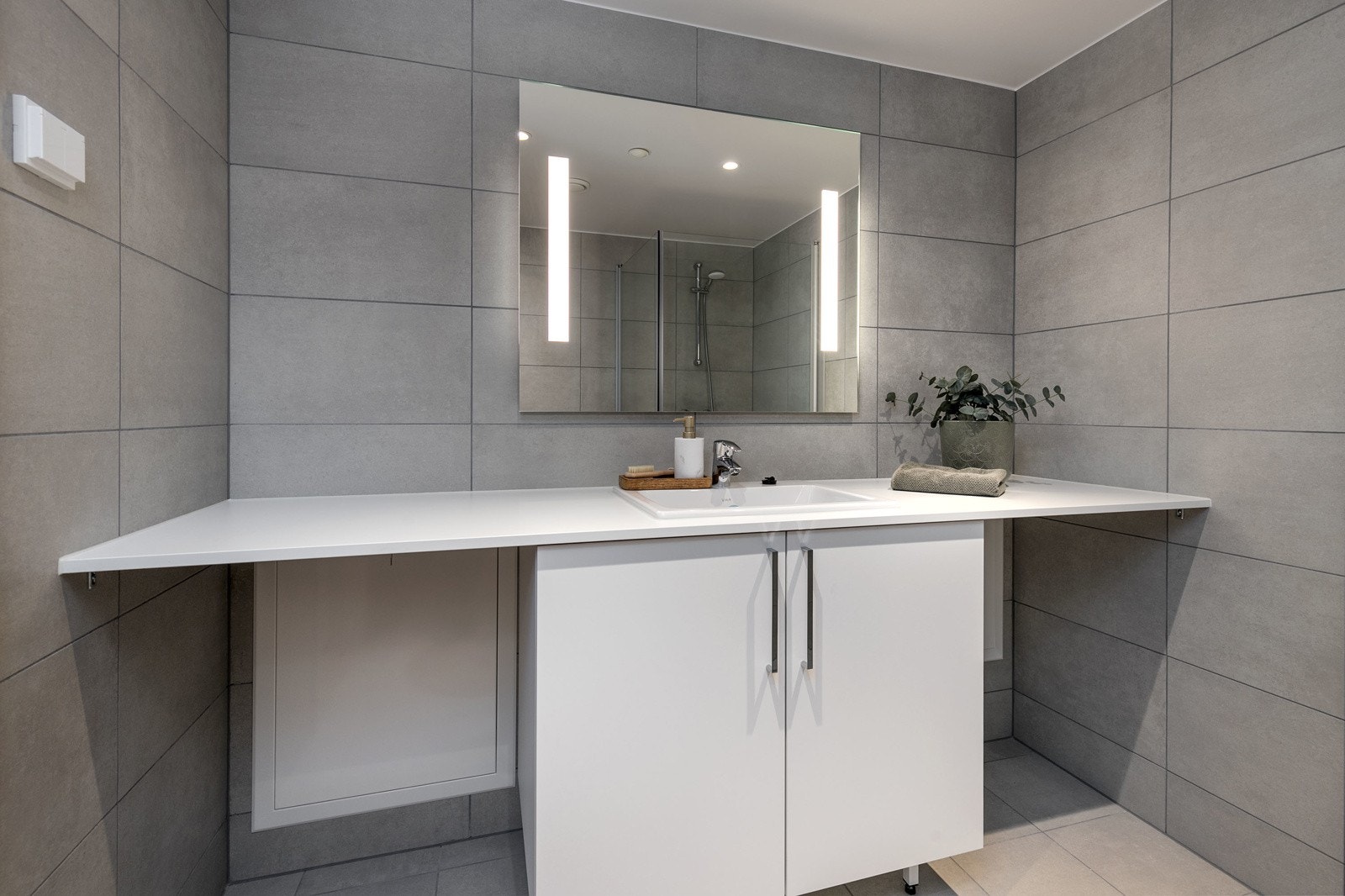 Delikat flislagt bad med vegghengt toalett, dusjsone med innfellbare dører samt servant med skapinnredning. Bilde fra L406. Galleribilde