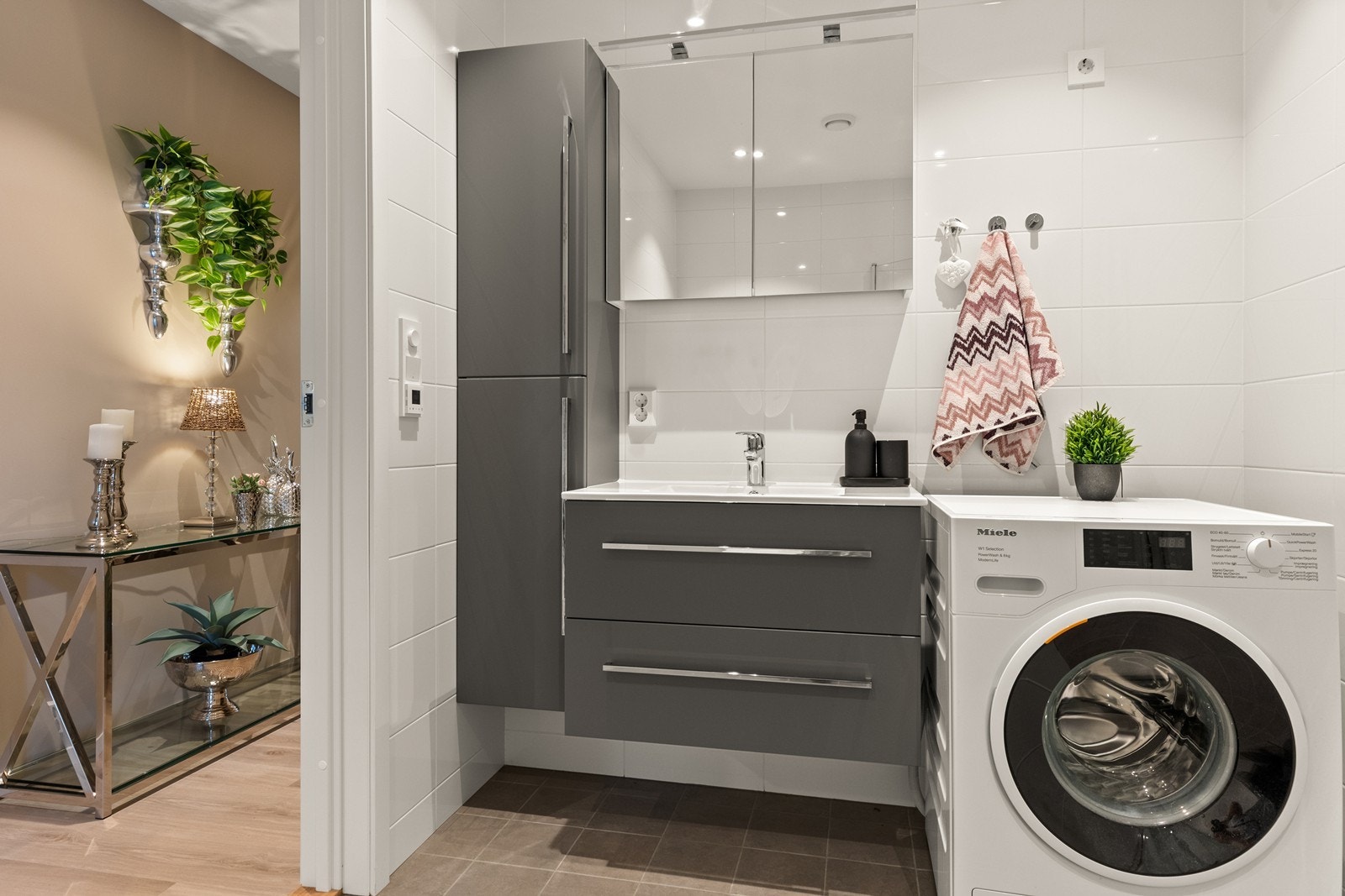 Bad i 1. etasje har grå gulvfliser og hvite veggfliser som gir et tidløst og rent uttrykk. Grå baderomsinnredning med skuffer og heldekkende servant, speilskap med belysning, samt et praktisk sideskap. Opplegg for vaskemaskin. Galleribilde