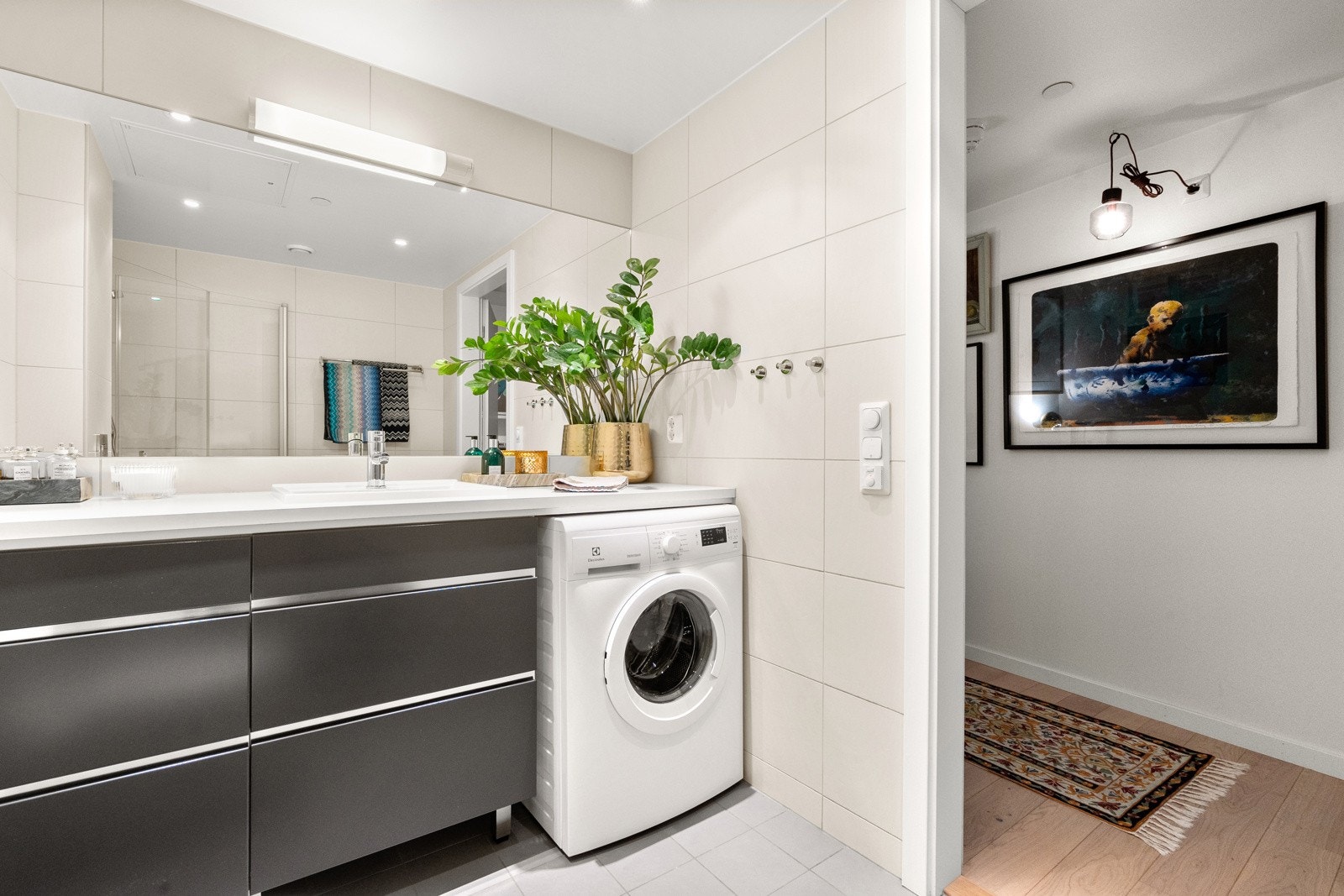 Moderne baderomsinnredning med skuffer og heldekkende benkeplate med nedsenket servant, stort speil med belysning over. Praktisk med plass og opplegg for vaskemaskin under benkeplaten. Galleribilde