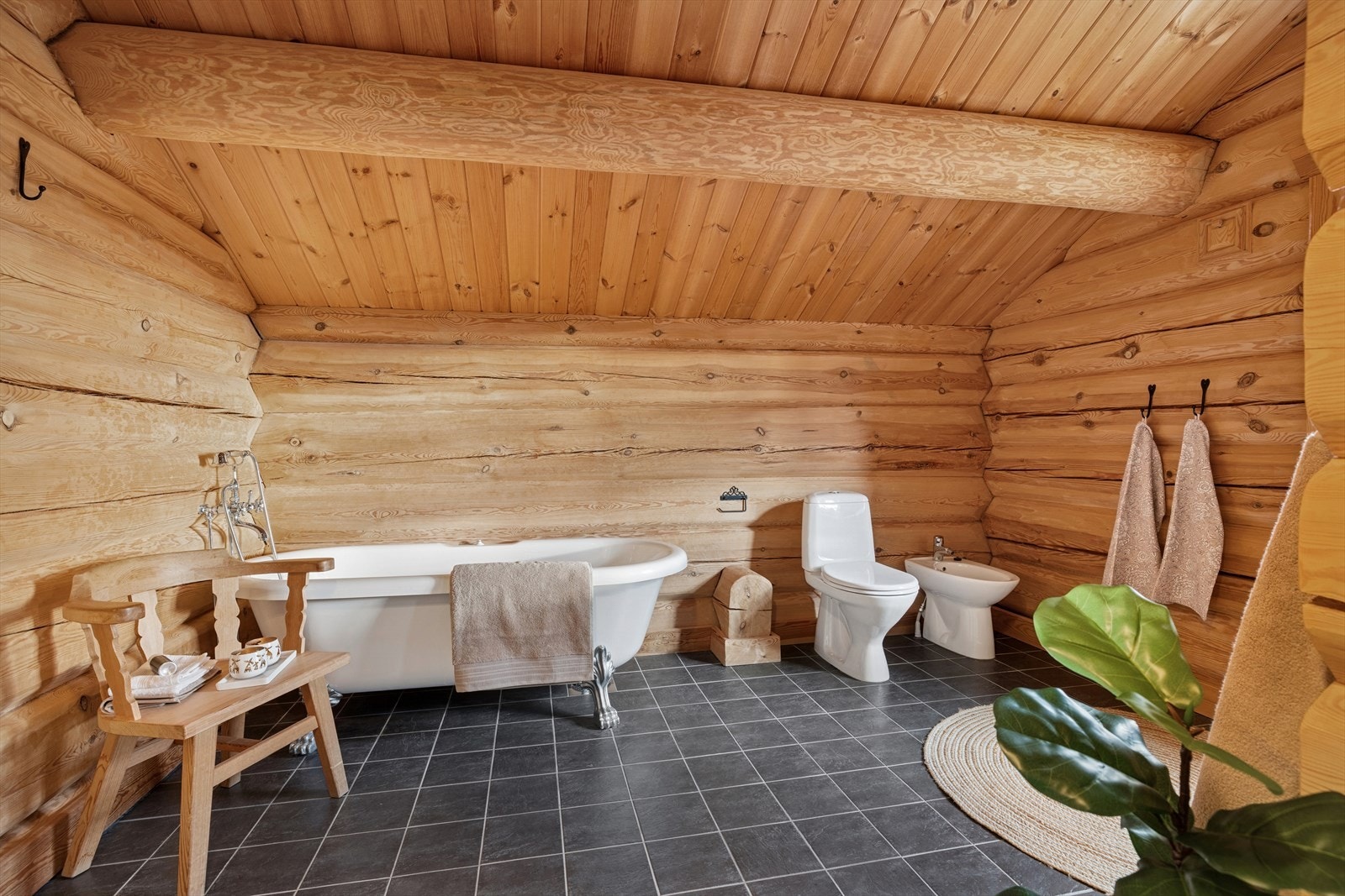 Dette badet kombinerer rustikk hyttestil med moderne komfort. De laftede veggene og synlige takbjelkene gir en varm og autentisk atmosfære, mens det flislagte gulvet med varmekabler sørger for praktisk bruk og behagelig temperatur. Galleribilde