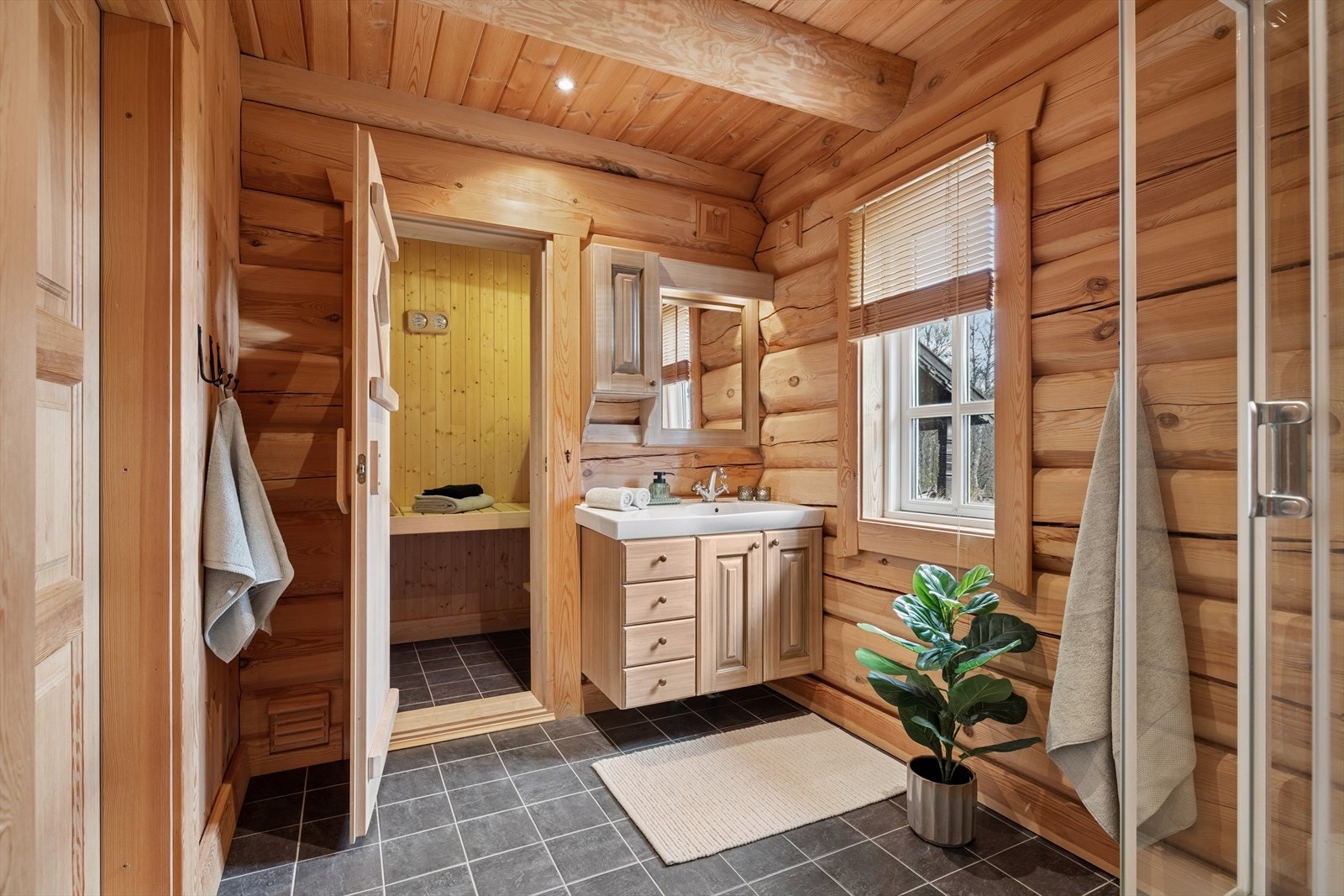 Badet i 1. etg. har en gjennomført stil med laftede vegger og varme tretoner som gir en lun og naturlig atmosfære. Rommet er romslig og har flislagt gulv med varmekabler for god komfort. Galleribilde