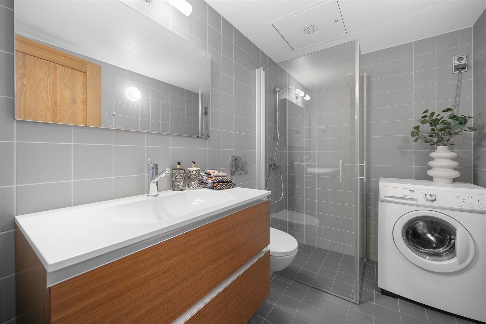 Baderom er bygd med flislagte gulv og vegger. Baderomsinnredning med heldekkende servant, ettgreps blandebatteri, dusjhjørne med glassdører og blandebatteri med temperaturinnstilling. Vegghengt toalett og opplegg for vaskemaskin. Galleribilde