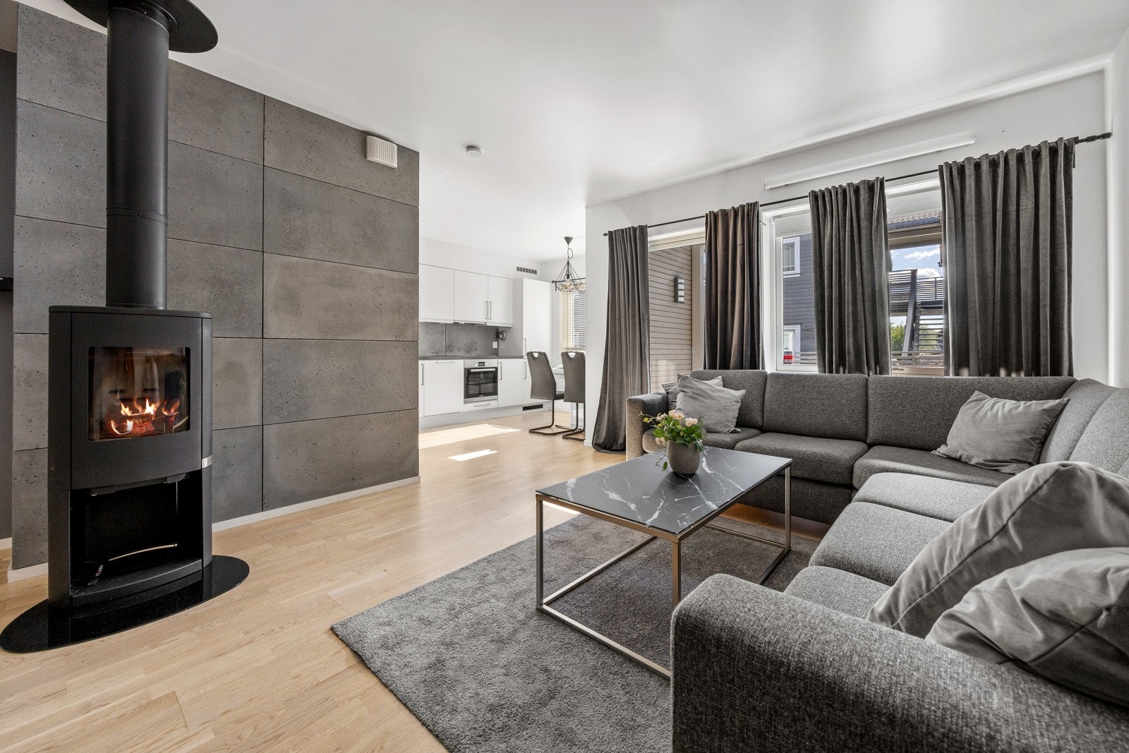 Midt i rommet er det en moderne peisovn som varmer godt på kalde dager og gir en lun atmosfære, varme og hygge. På gulvet er det lagt trestavs parkett som strekker seg sømløst inn i kjøkkenet. Galleribilde