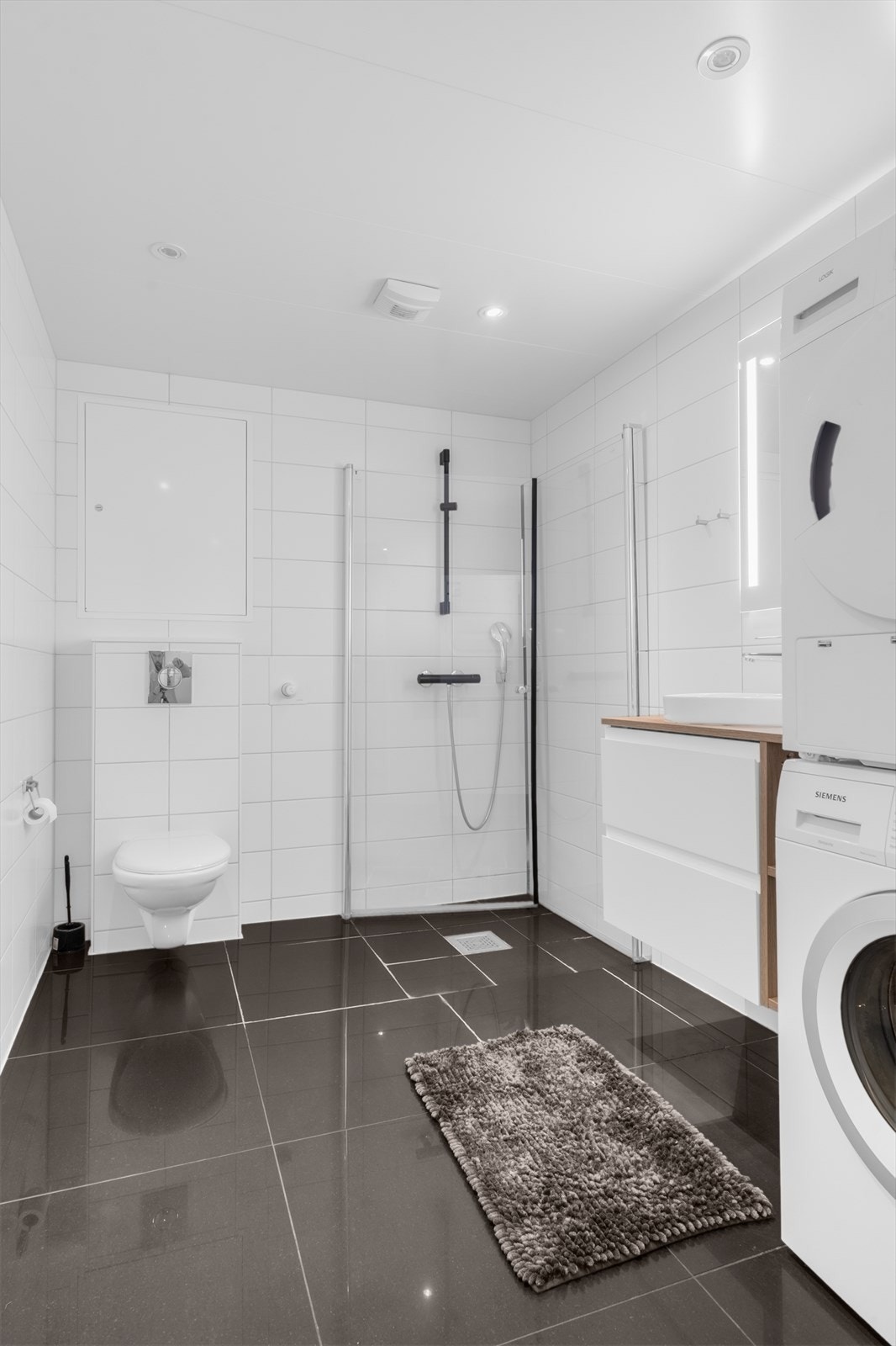 Flislagt baderom med innfelte dusjvegger i herdet glass, servant med underskap, speil med belysning og opplegg for vaskemaskin og tørketrommel. Galleribilde