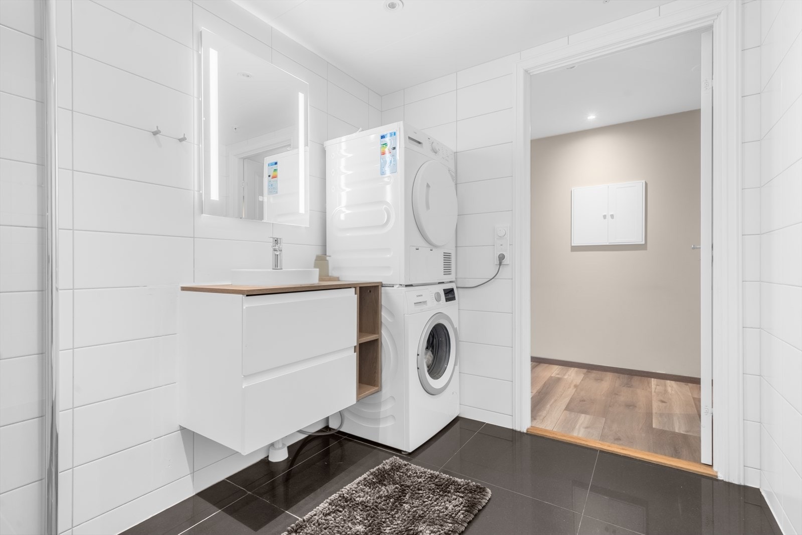 Flislagt bad med dusjhjørne, vegghengt toalett og opplegg for vaskemaskin og tørketrommel. Galleribilde