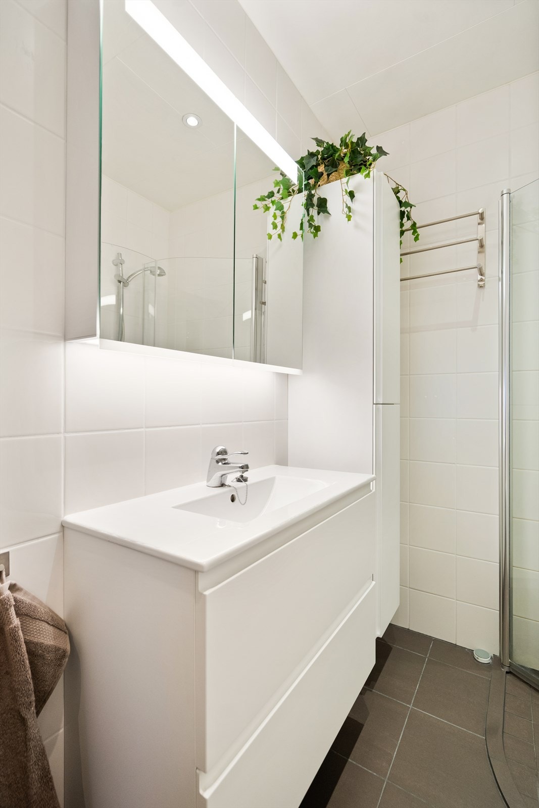 Badet er utstyrt med dusjhjørne, veggmontert toalett, baderomsinnredning med underskap, høyskap og overskap med lys og speil. Galleribilde