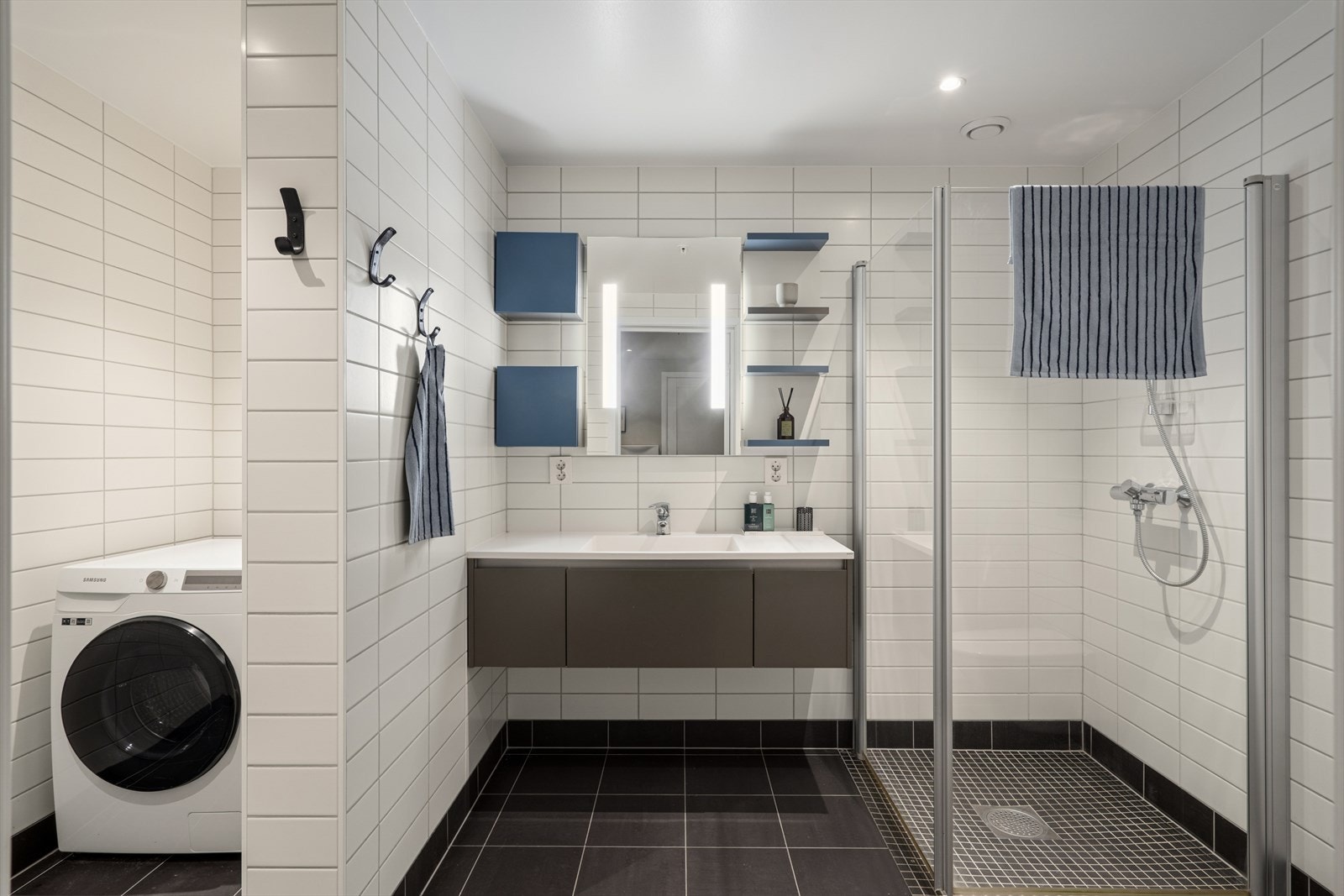 Bad/vaskerom er innredet med servant på skuffeseksjon, speil med integrert lys, vegghengt toalett og et dusjhjørne med glassdører. Det er også opplegg for vaskemaskin. Galleribilde