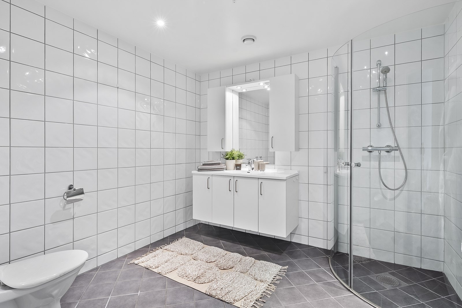 Badet er utstyrt med heldekkende servant over skapinnredning, speil med overliggende belysning, vegghengt skap. dusjhjørne med innfellbare glassvegger og toalett Galleribilde