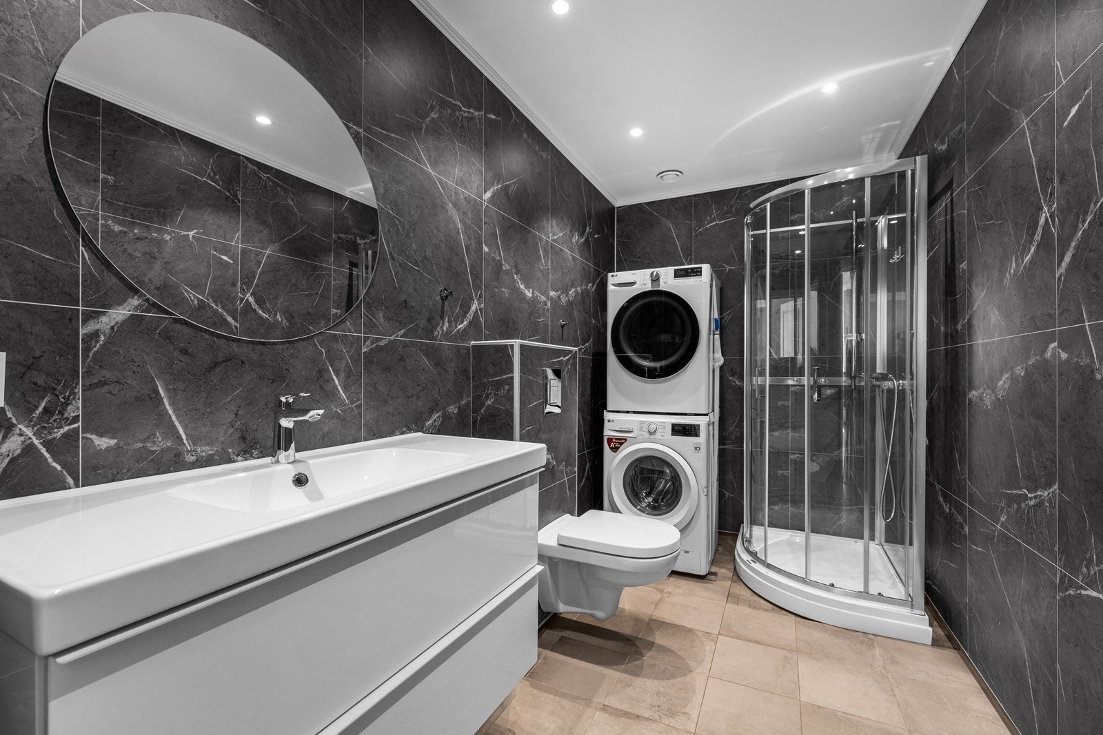 Pent baderom innredet med helservant på skuffeseksjon, vegghengt toalett, dusjkabinett og opplegg for vaskemaskin. Galleribilde