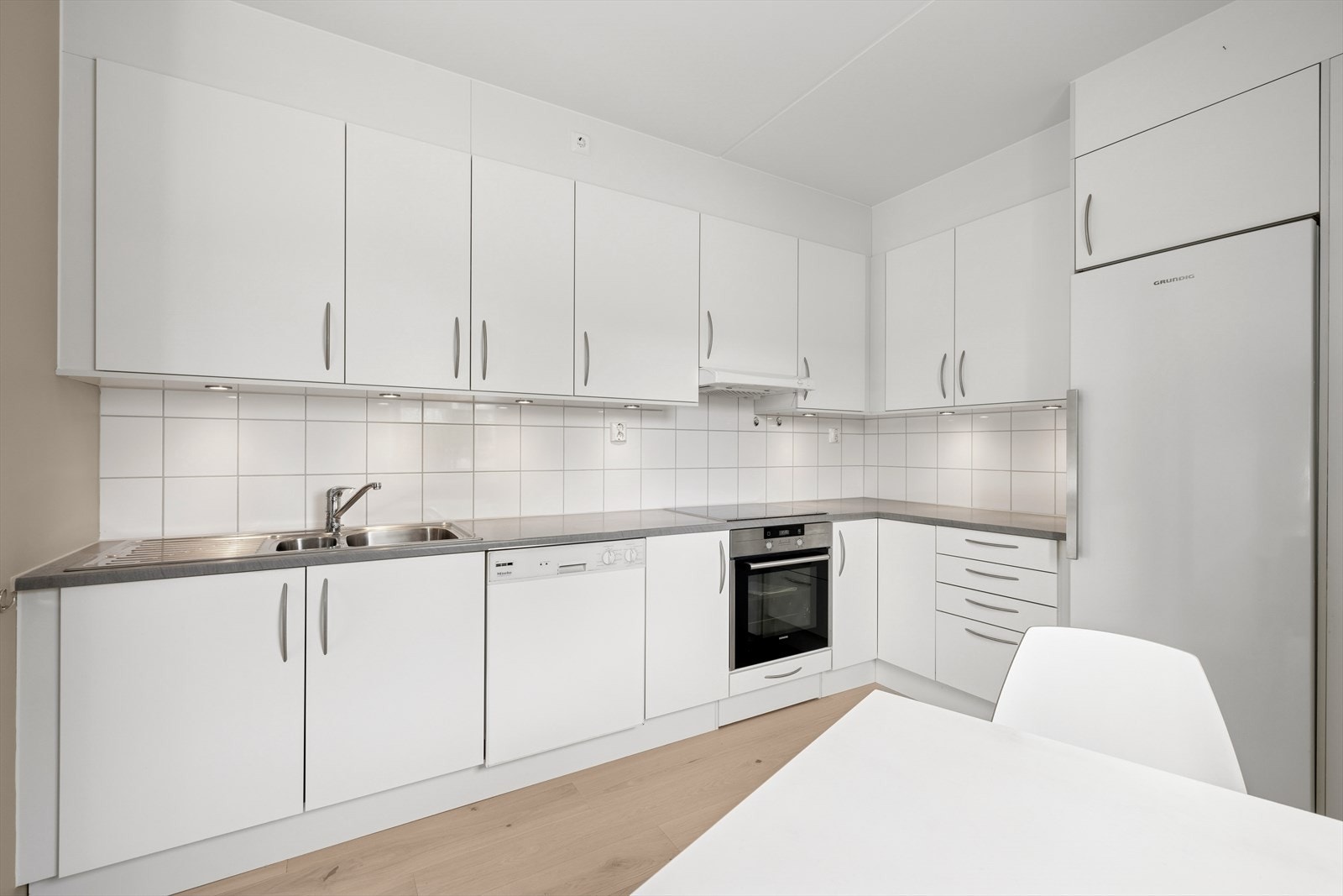 Integrert Induksjonstopp og stekeovn, frittstående kjøleskap og oppvaskmaskin. Det er keramiske fliser mellom overskap og benkeplate. Galleribilde