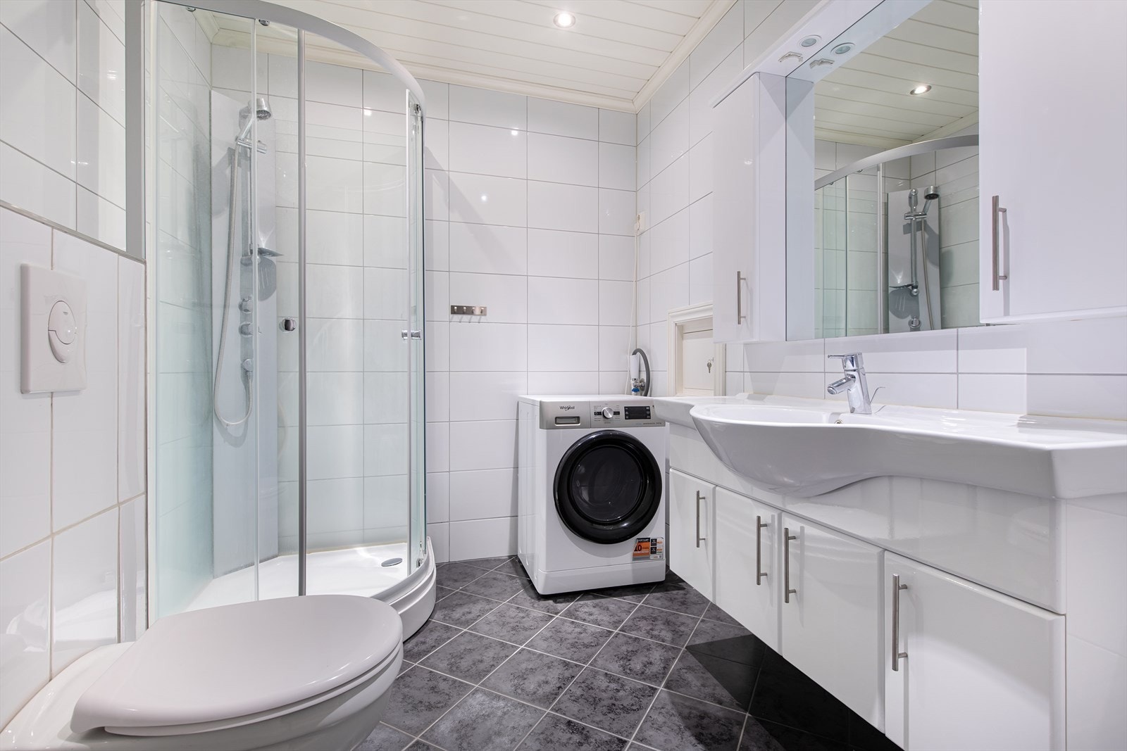 Bad/wc i 1 etg med tilkomst fra stue/kjøkken. I rommet er det vaskeservant i innredning, veggmontert wc, lukket dusjkabinett og opplegg for vaskemaskin. Galleribilde