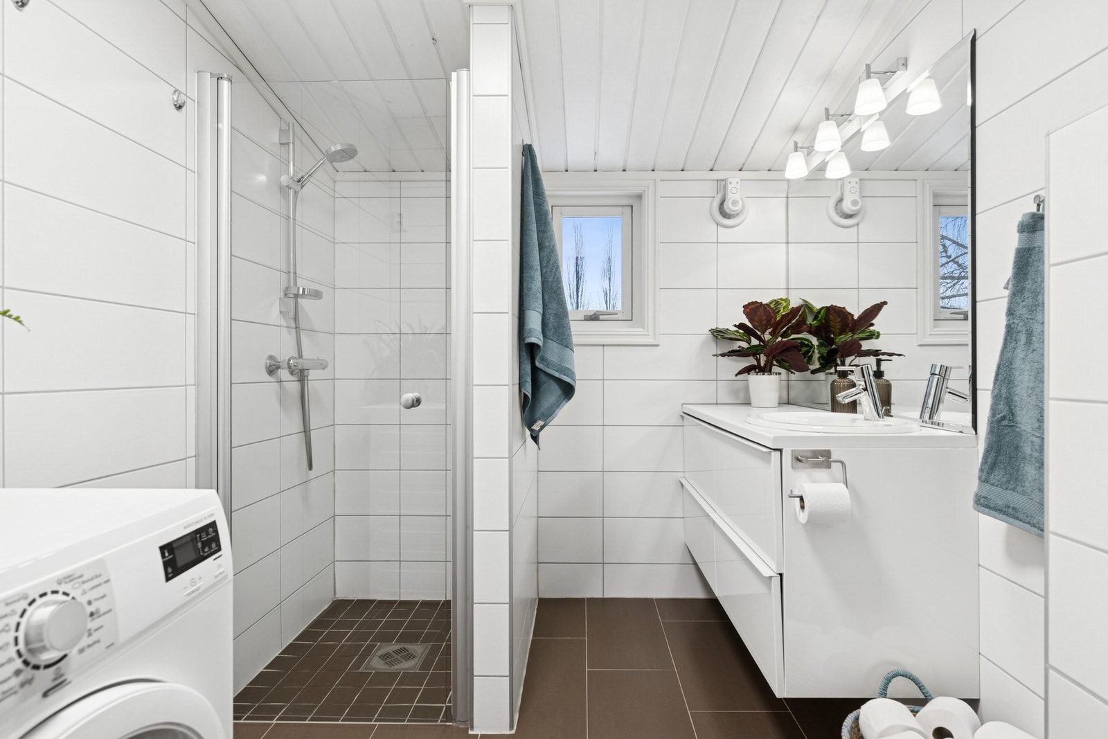 Baderom med fliser på gulv og vegger, hvit servantinnredning med skuffeseksjon, nedsenket dusjnisje med glassdør og vegghengt toalett. Galleribilde