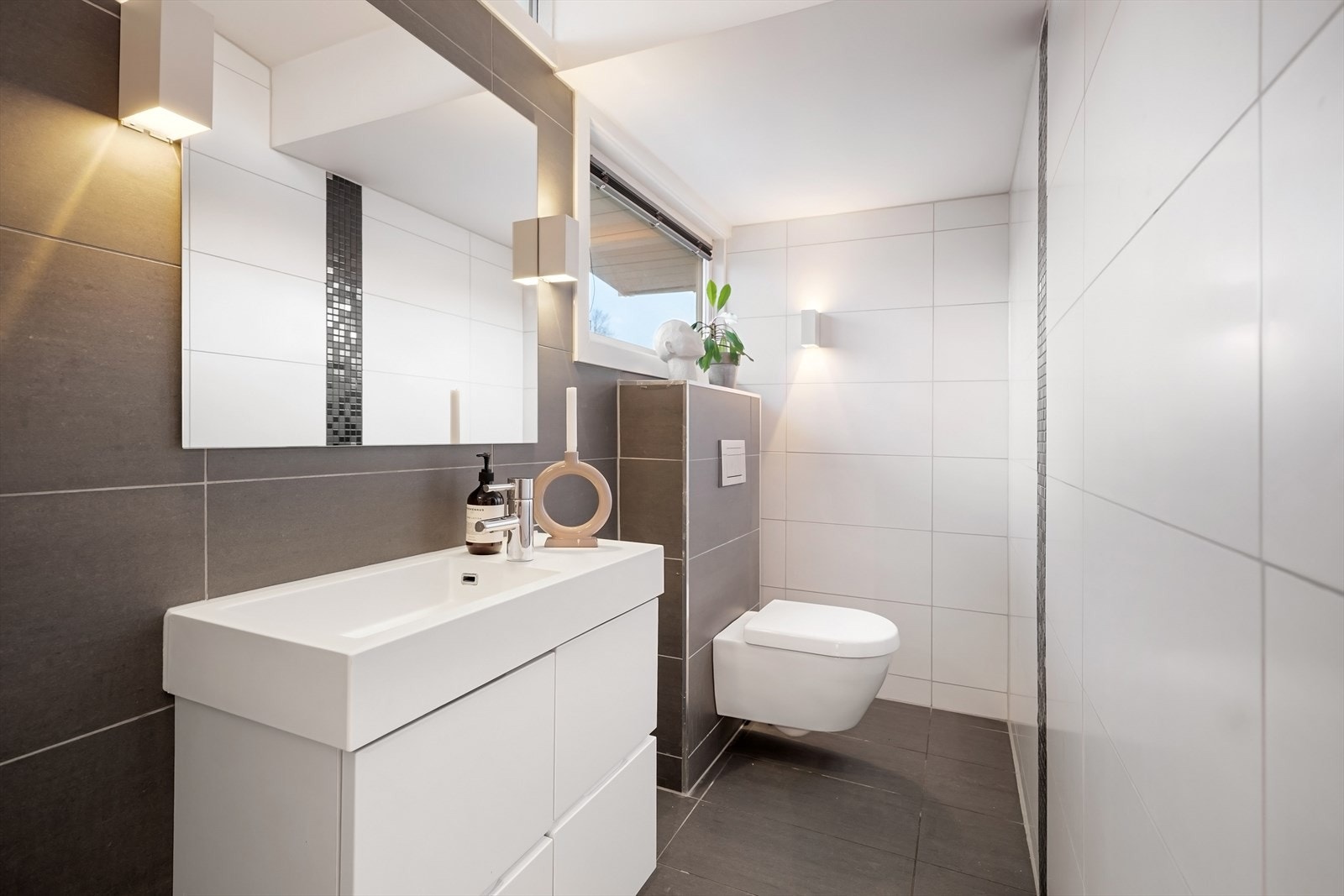 Fra entréen er det tilgang til et praktisk toalettrom med fliselagte overflater. Vegghengt wc og baderomsinnredning med skap og heldekkende servant med ettgreps blandebatteri. Vegglamper fungerer som lyskilde, i tillegg til naturlig lysinnslipp fra vindu. Galleribilde