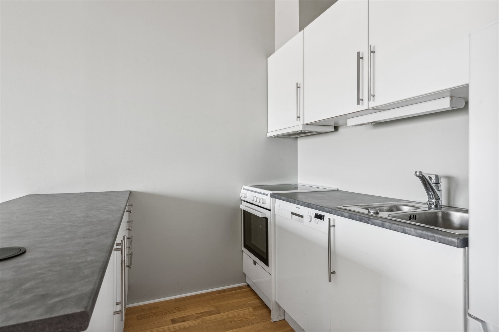 Innredningen har hvite, slette fronter kombinert med grå laminat benkeplate. Hvitevarer består av komfyr og innebygget oppvaskmaskin. Galleribilde