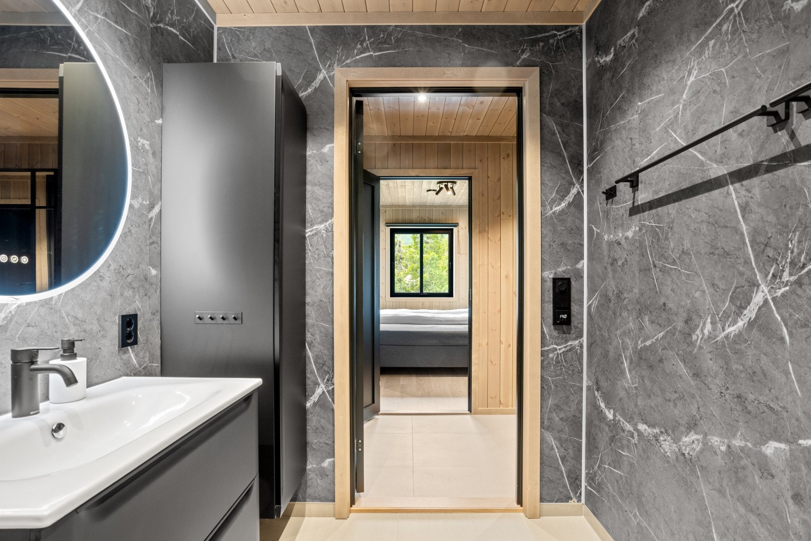 Baderomsinnredningen består av et heldekkende baderomsservant med skuffer og et høyskap, begge med slette fronter i dempet sort. Over servantløsningen henger et moderne, rundt smartspeil med integrert belysning og avdamping. Galleribilde