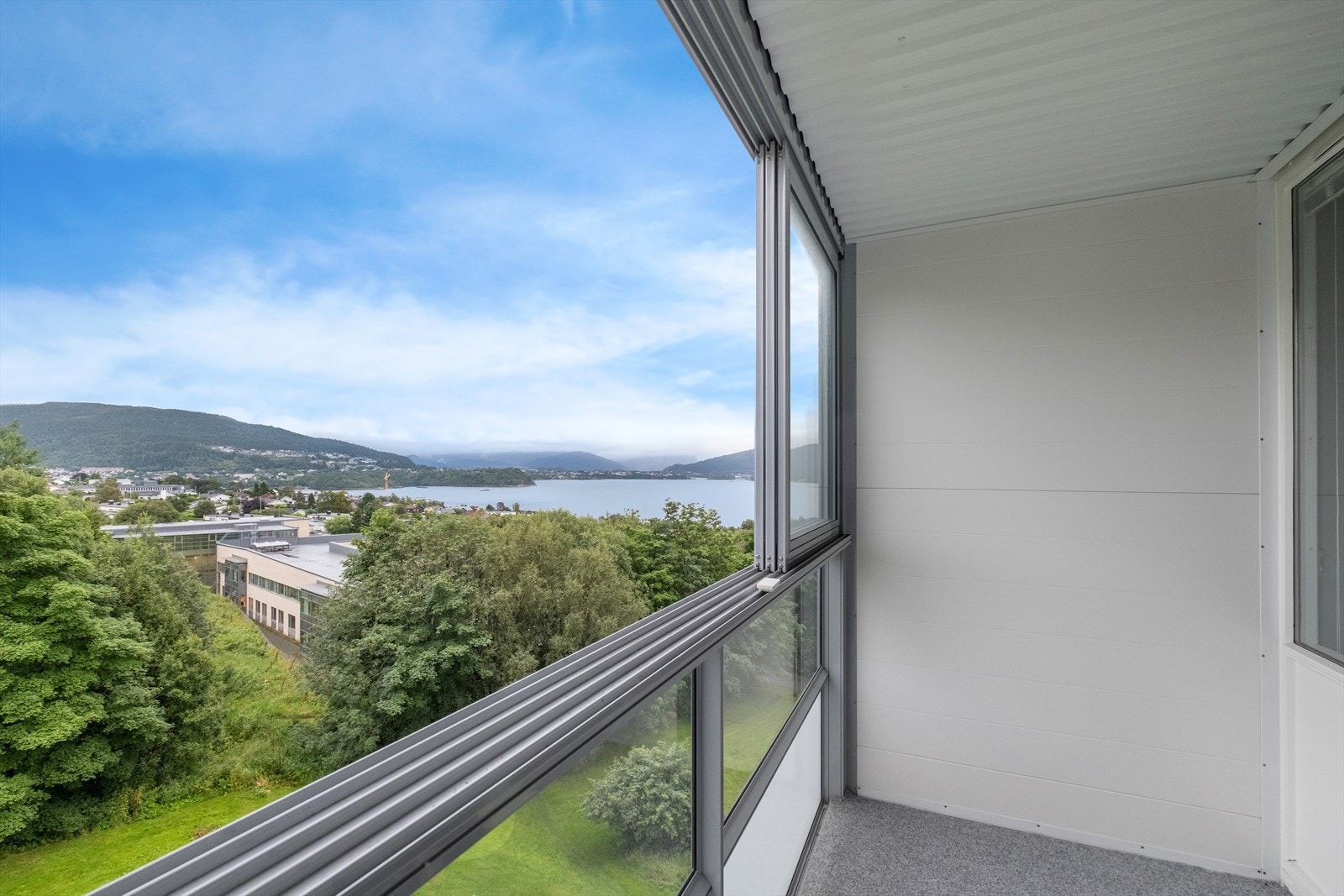 Innglasset balkong med teppebelagt betongdekke gir utsikt mot sjøen og grøntområdet. Galleribilde