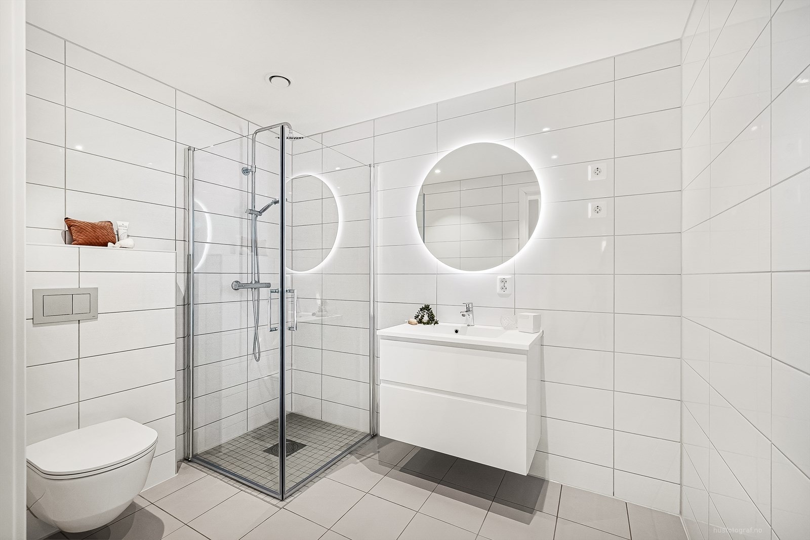 Lekkert flislagt baderom med vegghengt toalett, dusjnisje, skuffeinnredning med nedfelt vask og rundt speile med innebygd lys. Galleribilde