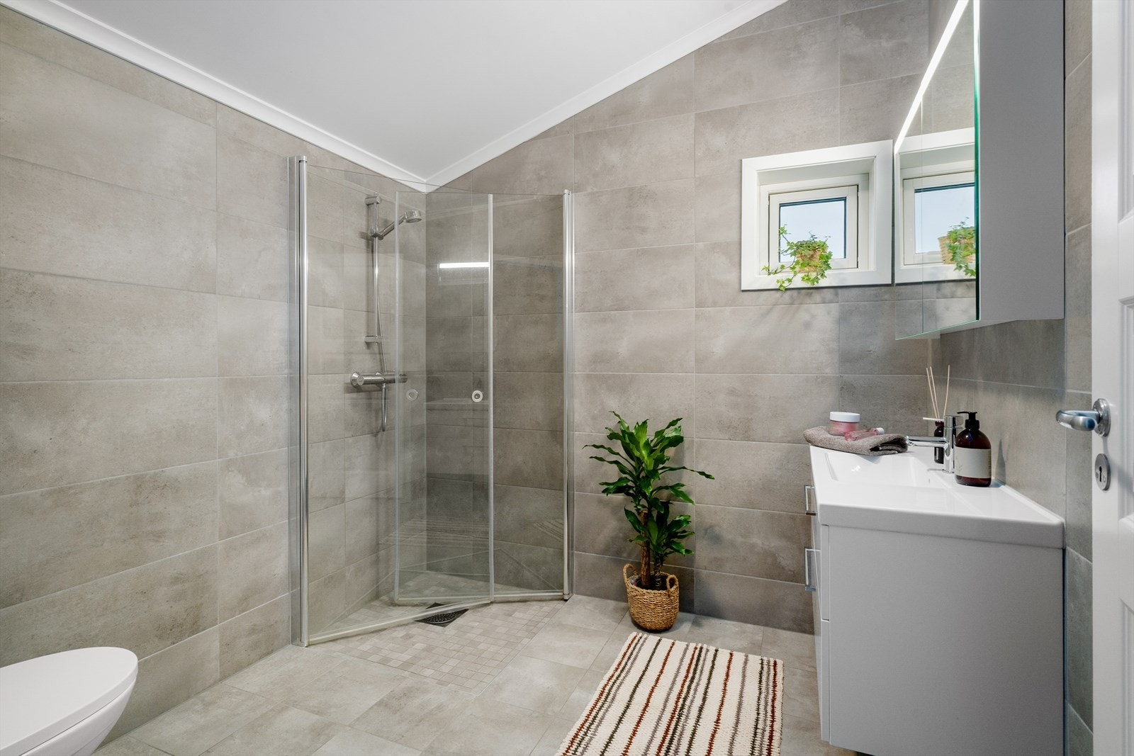 Lekkert bad med flis på gulv og vegger. Badet har vegghegghengt toalett, dusjhjørne med glassdører og garnityr, samt innredning med heldekkende hvit vask, underskuffer med slette fronter og overskap med speildører. Galleribilde