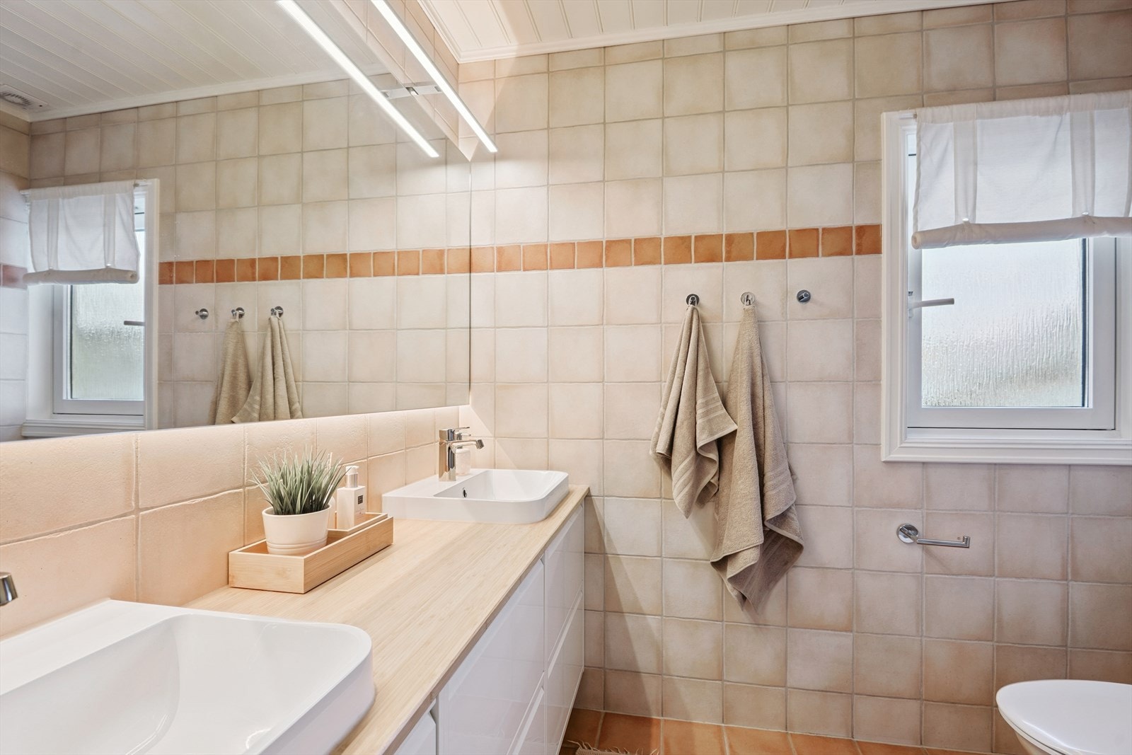 Flislagt bad med elektriske varmekabler i gulv. Innredningen består av to nedfelte servanter, gulvstående toalett og et dusjkabinett. Galleribilde