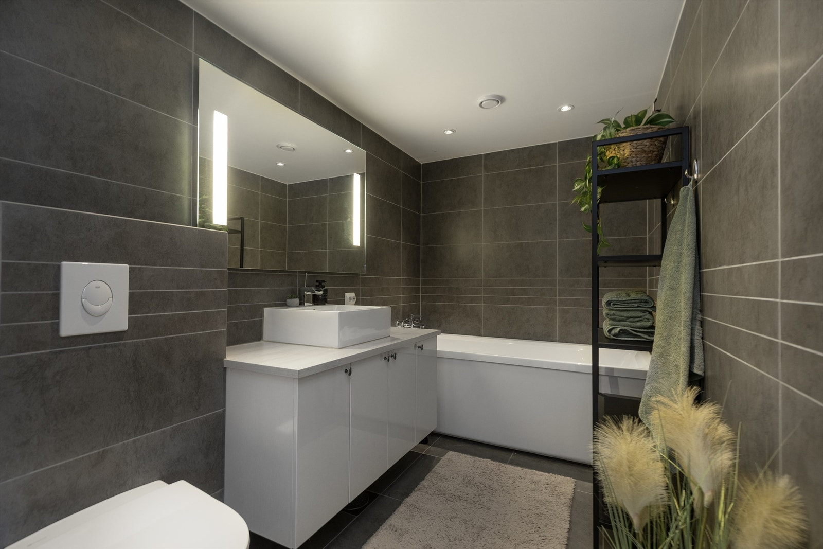 Moderne flislagt bad med mørke fliser, stilrene overflater og funksjonell innredning. Badet tilbyr blant annet vannbåren gulvvarme, stemningsfull belysning med downlights. Galleribilde