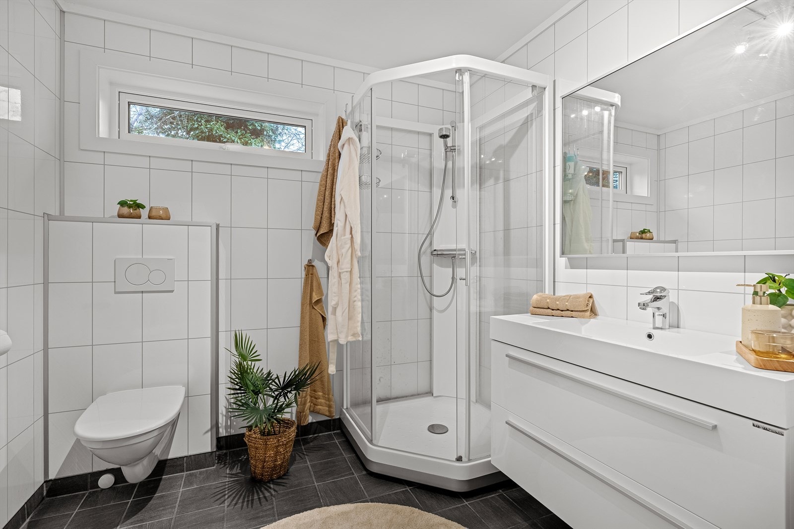 Badet er lyst og trivelig med fliser på gulver hvor det er varmekabler. Det inneholder ellers et vegghengt toalett, dusjkabinett, samt baderomsinnredning med speil over. Det er også opplegg og plass til vaskemaskin/tørketrommel. Galleribilde